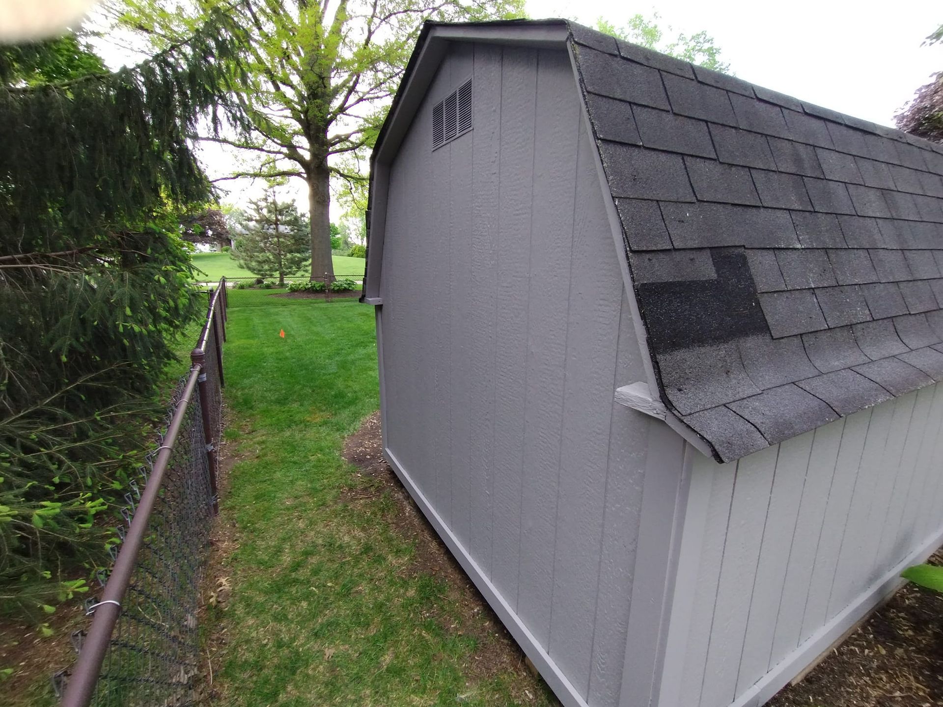 A gray shed with a black roof is next to a chain link fence.