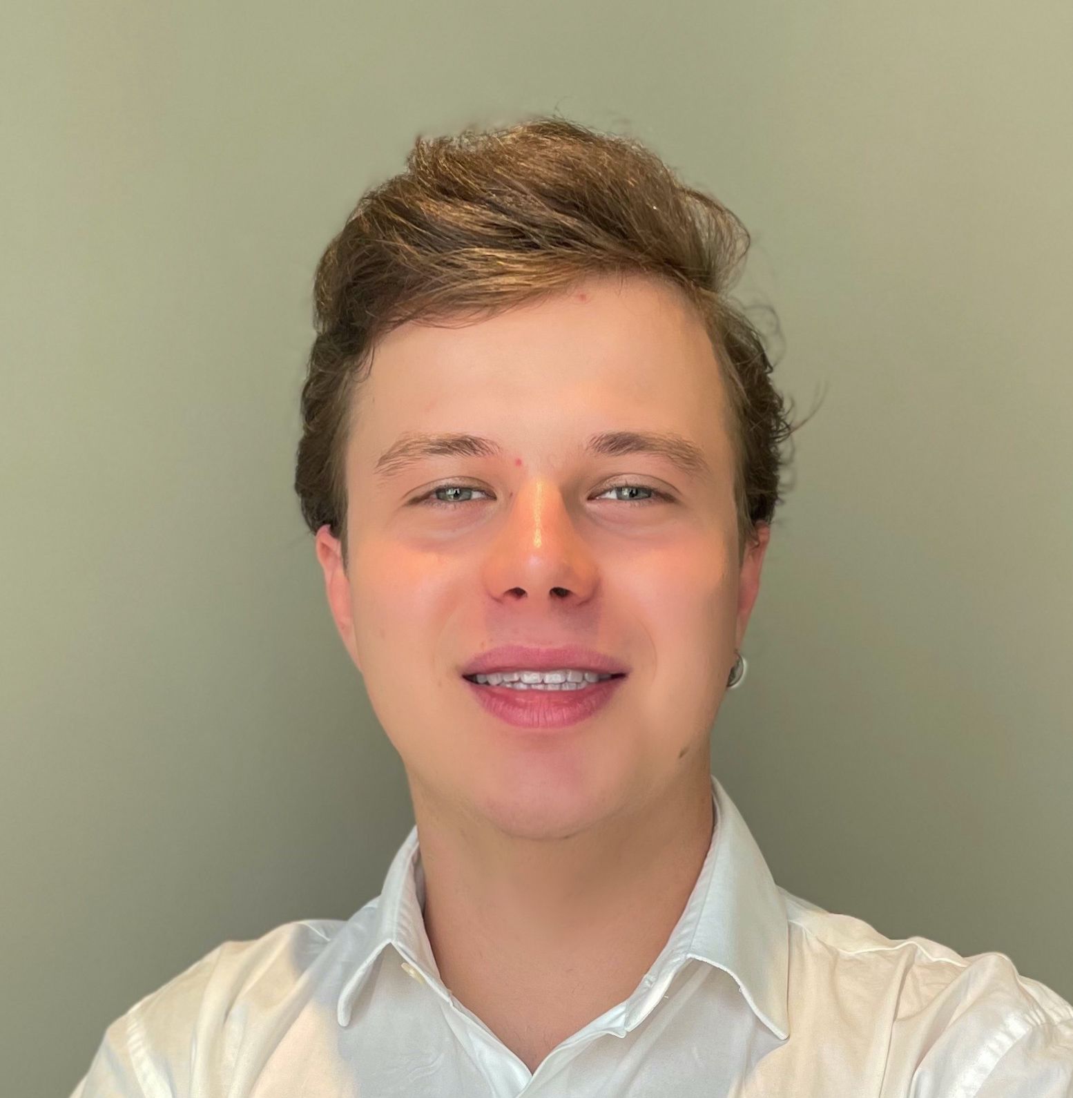 Homem de pele clara e cabelo castanho sorrindo contra um fundo verde, vestindo uma camisa branca.