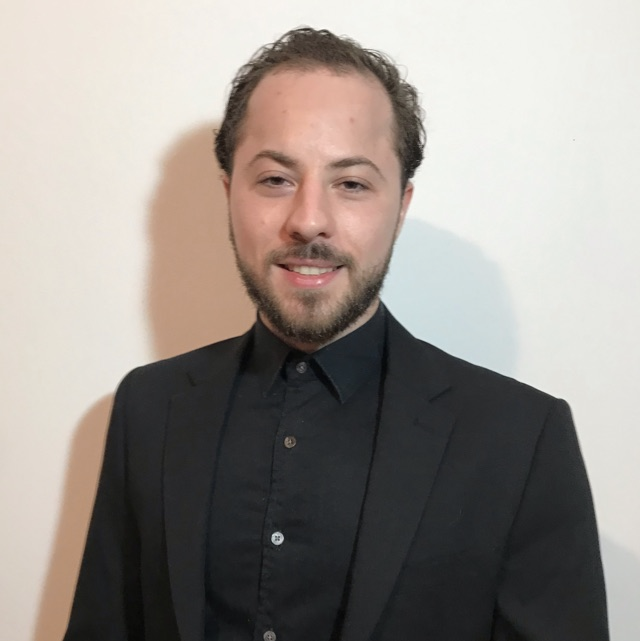 Homem com cabelo castanho curto e barba, vestindo camisa preta e paletó, sorrindo para a câmera.