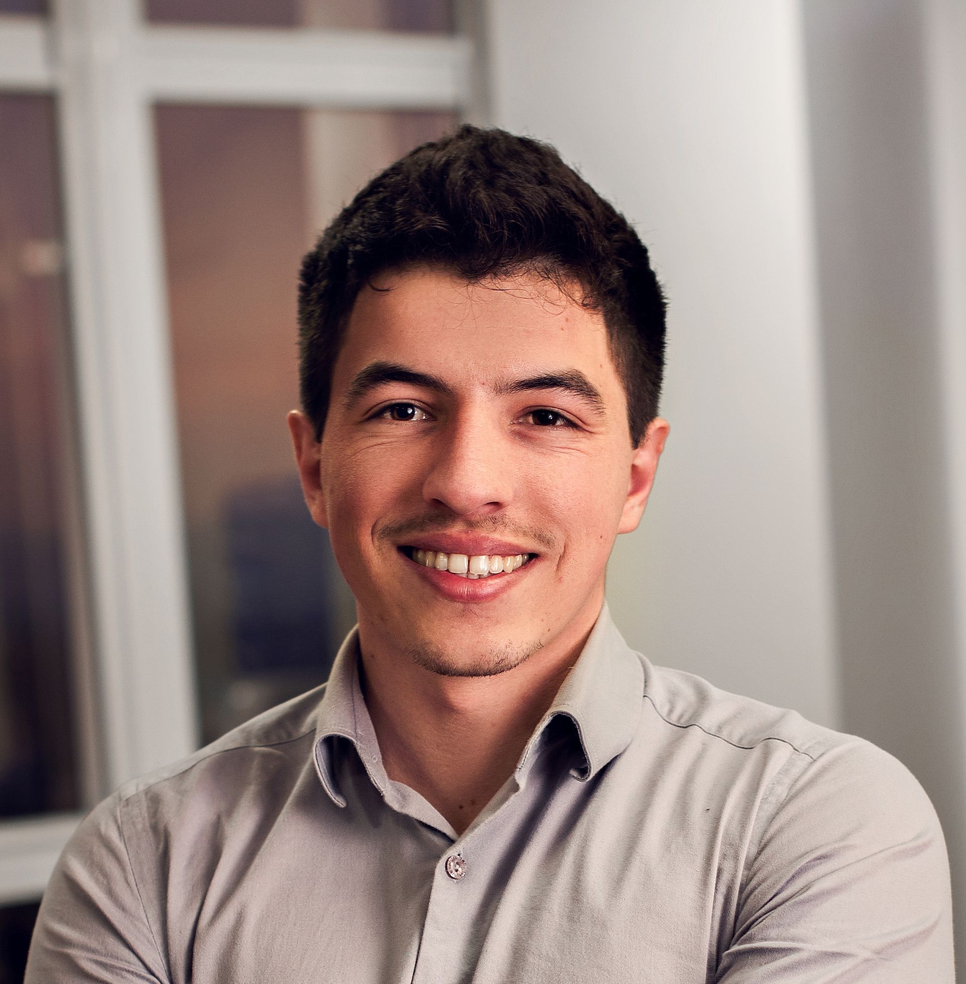 Homem sorridente vestindo uma camisa cinza de botões, em ambiente interno.