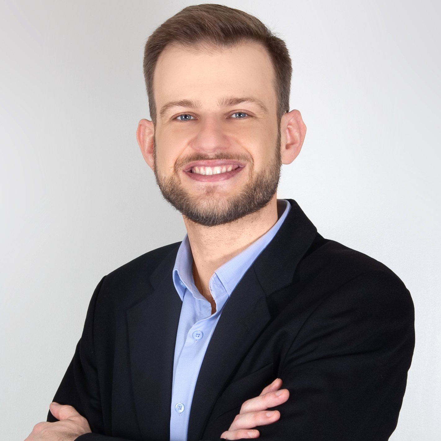 Homem barbudo, vestindo blazer preto e camisa azul, sorri com os braços cruzados.