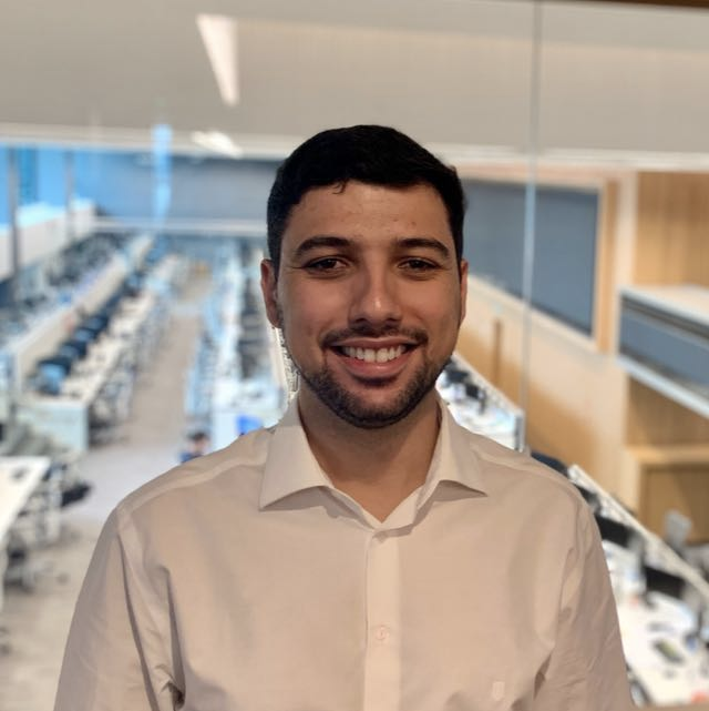 Homem de camisa branca sorrindo em um escritório com fileiras de mesas.
