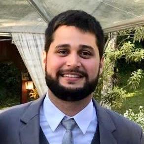 Um homem de terno cinza sorri ao ar livre, perto de um prédio com cortinas e folhagem.