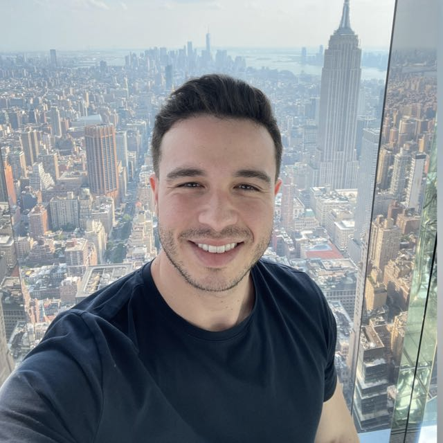 Homem sorrindo enquanto tira uma selfie com vista para a paisagem urbana de Nova York, incluindo o Empire State Building.