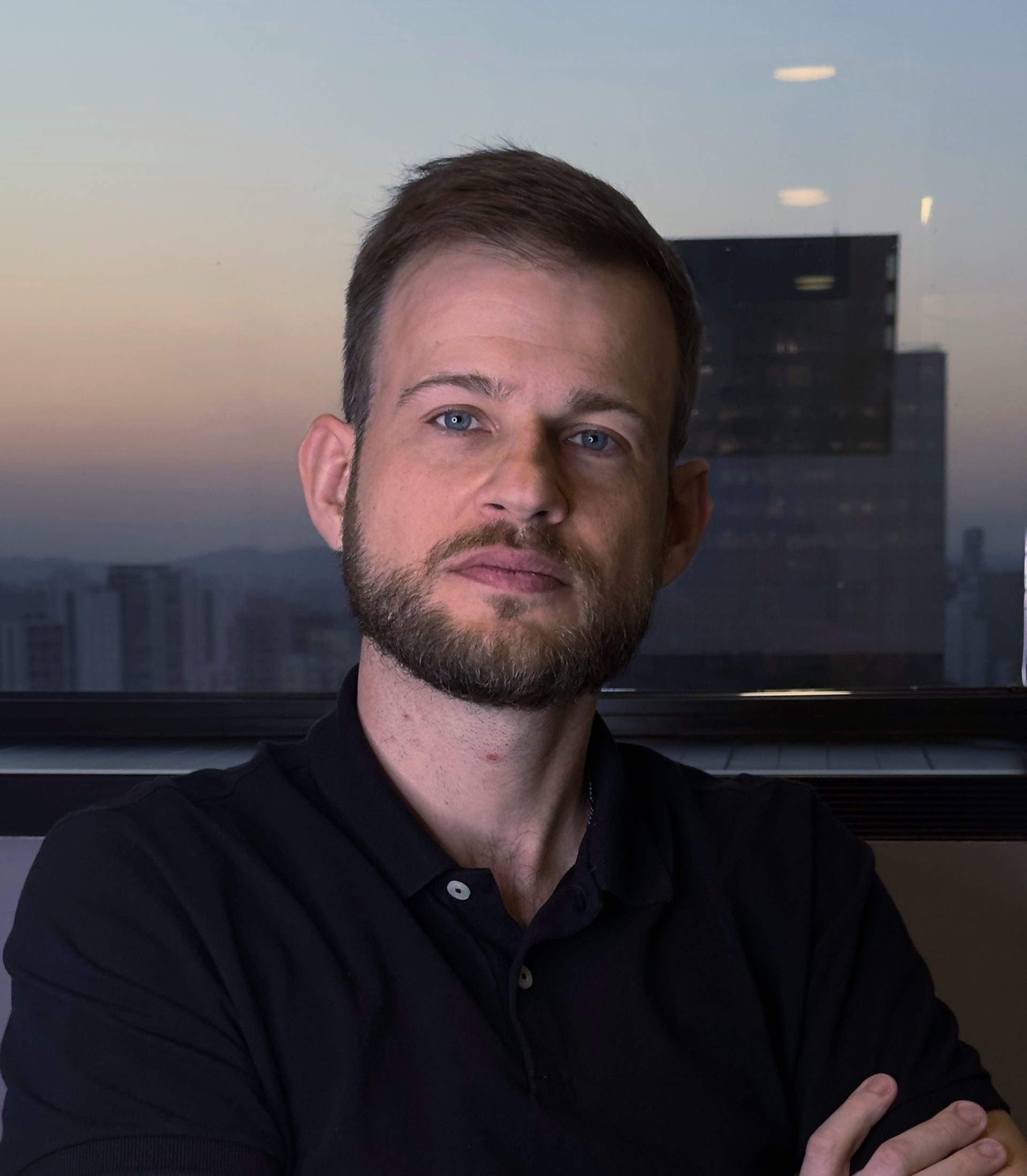 Homem com barba curta, vestindo camisa preta, olhando para a câmera em um ambiente de escritório.