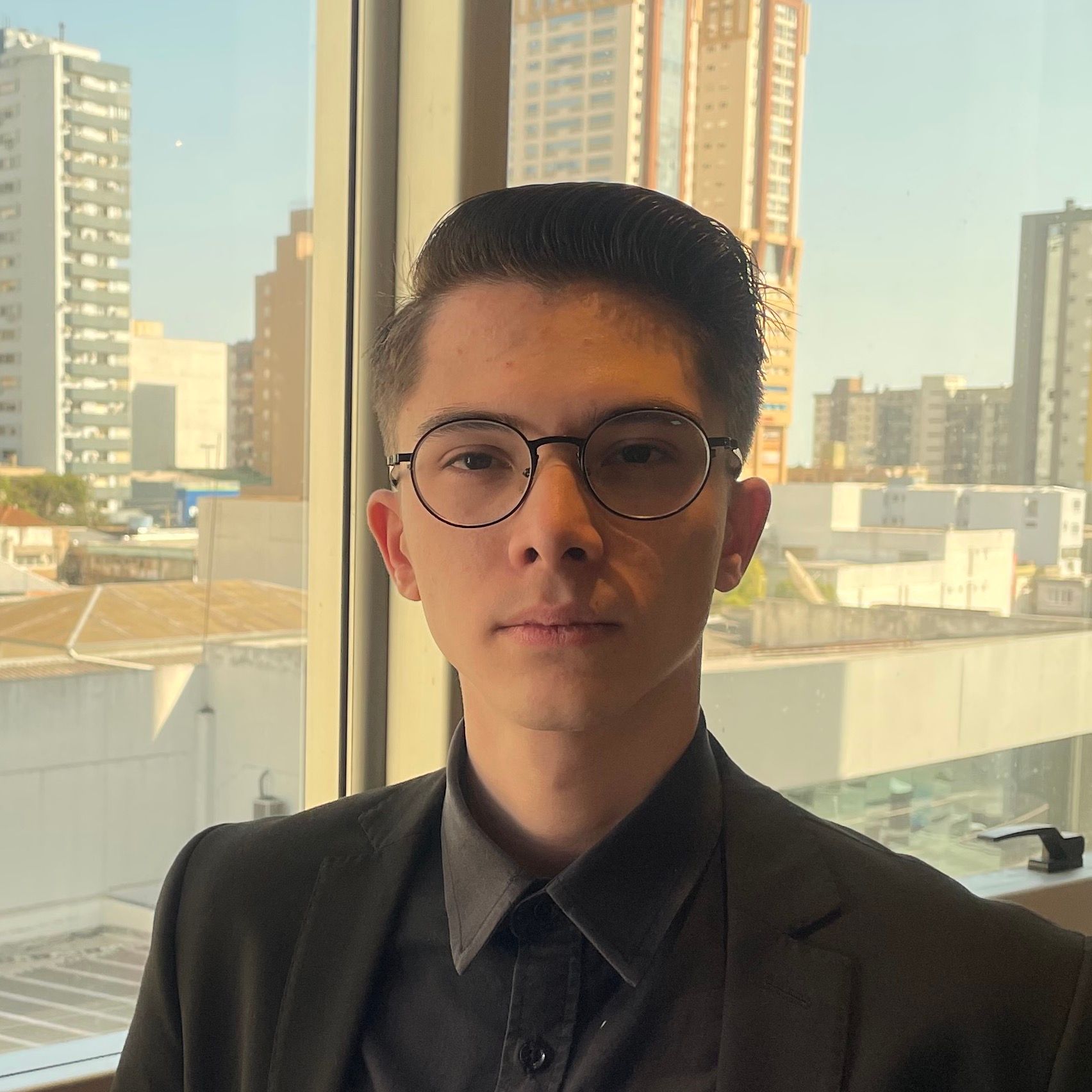 Homem de terno preto e óculos, olhando para a câmera, com paisagem urbana ao fundo.