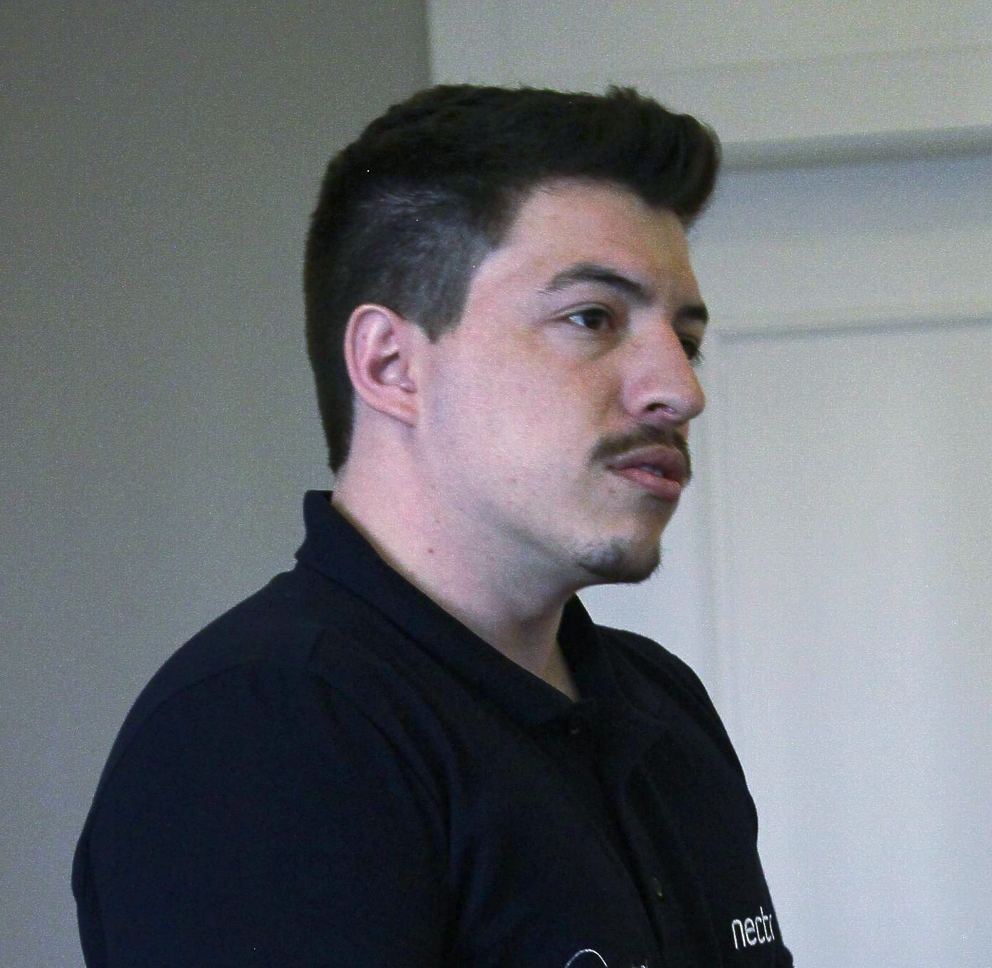 Homem de camisa escura olhando para a direita, em um ambiente claro. Ele tem bigode e cabelo curto e escuro.