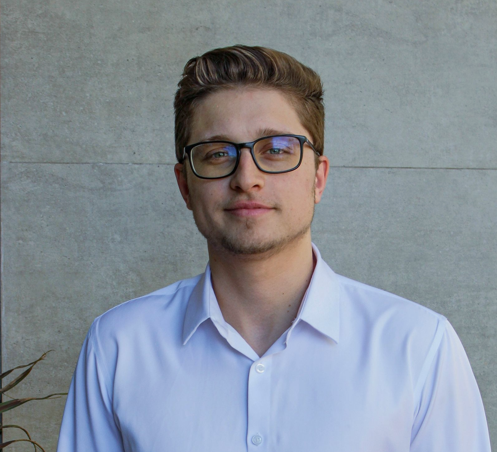 Um homem de óculos e camisa branca de botões sorri, em pé em frente a uma parede de cor neutra.