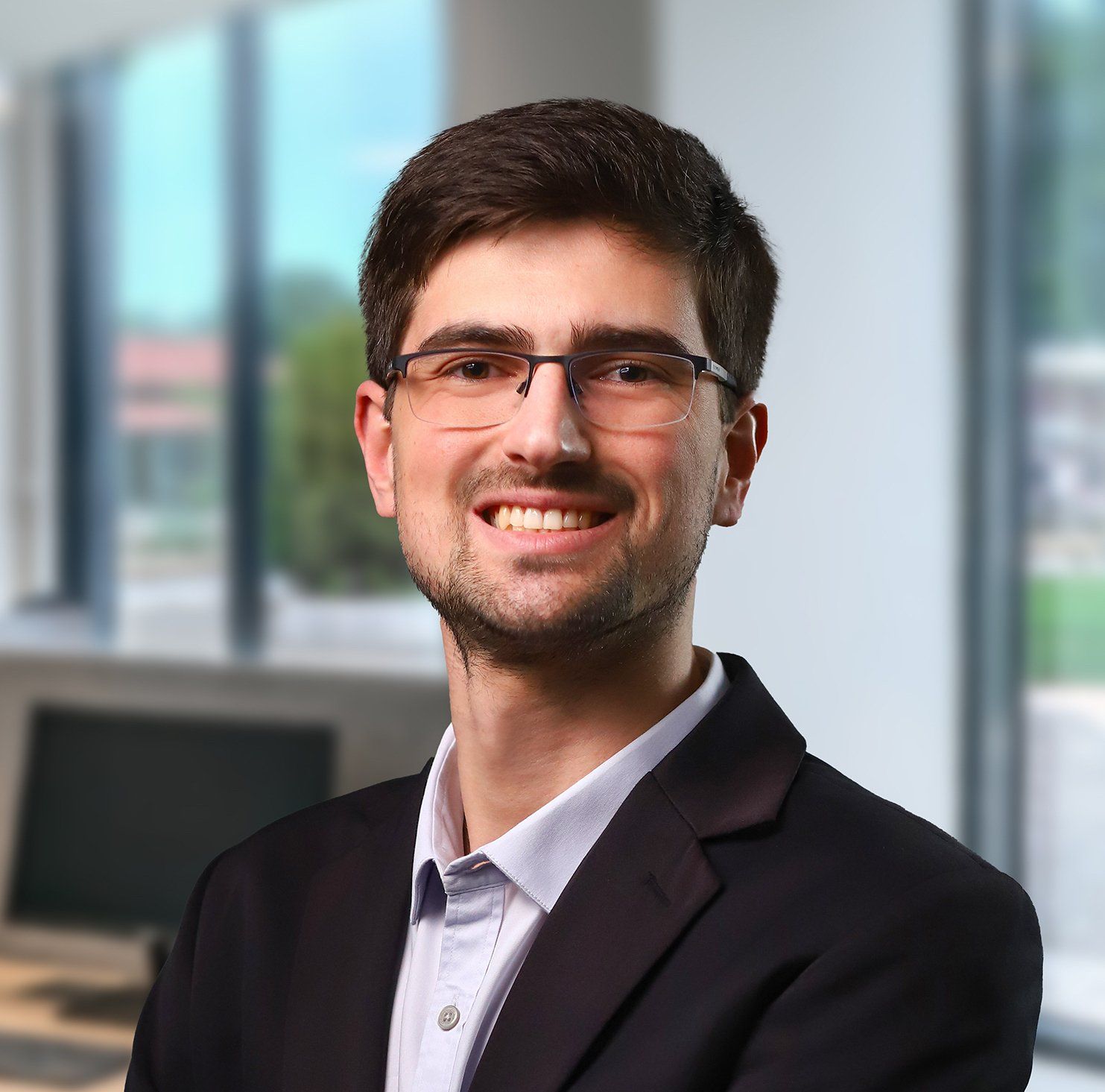 Homem de óculos sorrindo, vestindo paletó e camisa de colarinho em um ambiente de escritório.