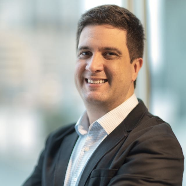 Homem de blazer e camisa com a gola aberta, sorrindo, em ambiente interno com fundo desfocado.