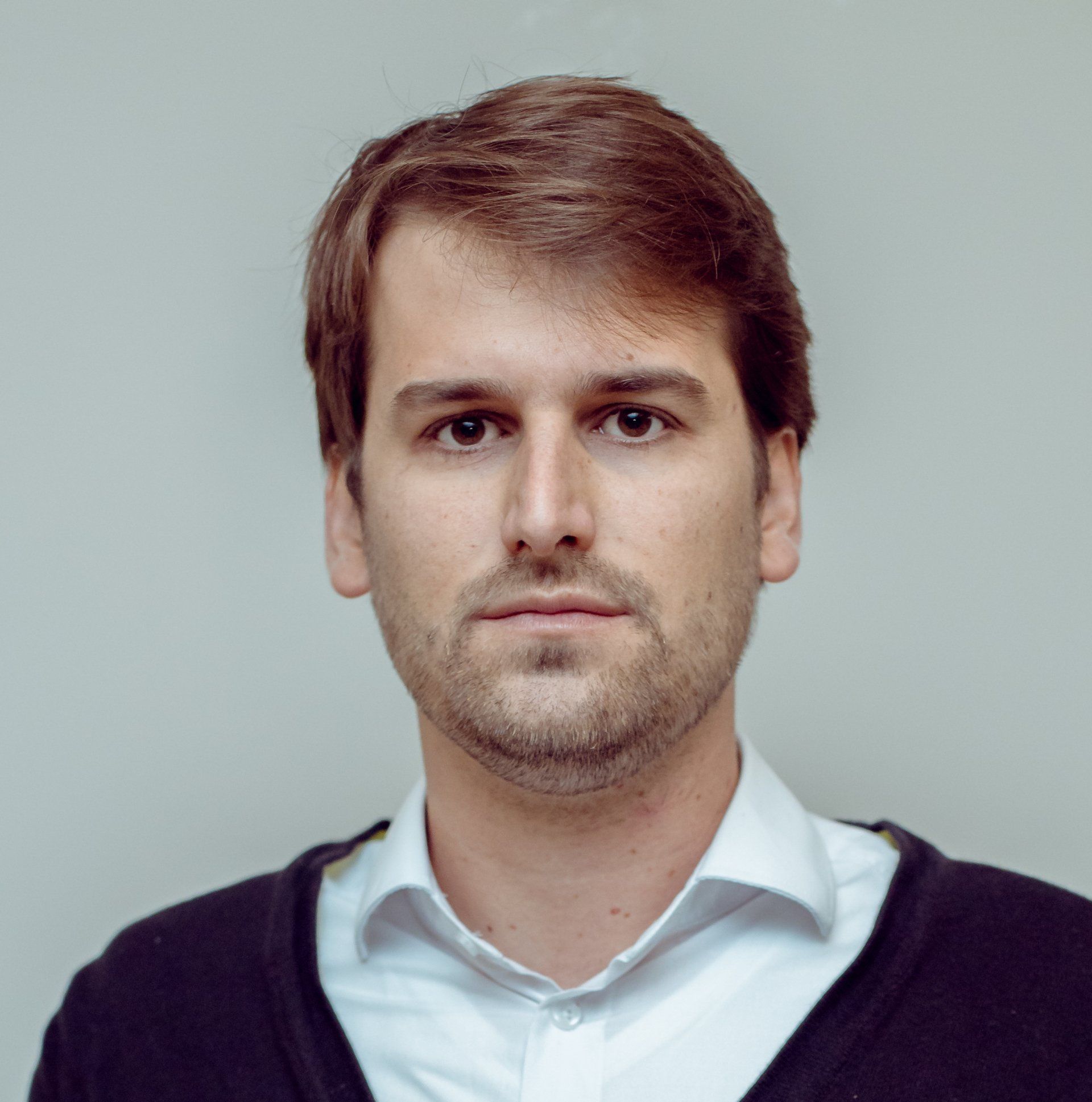 Homem de camisa branca e suéter escuro, olhando para a câmera. Expressão neutra.