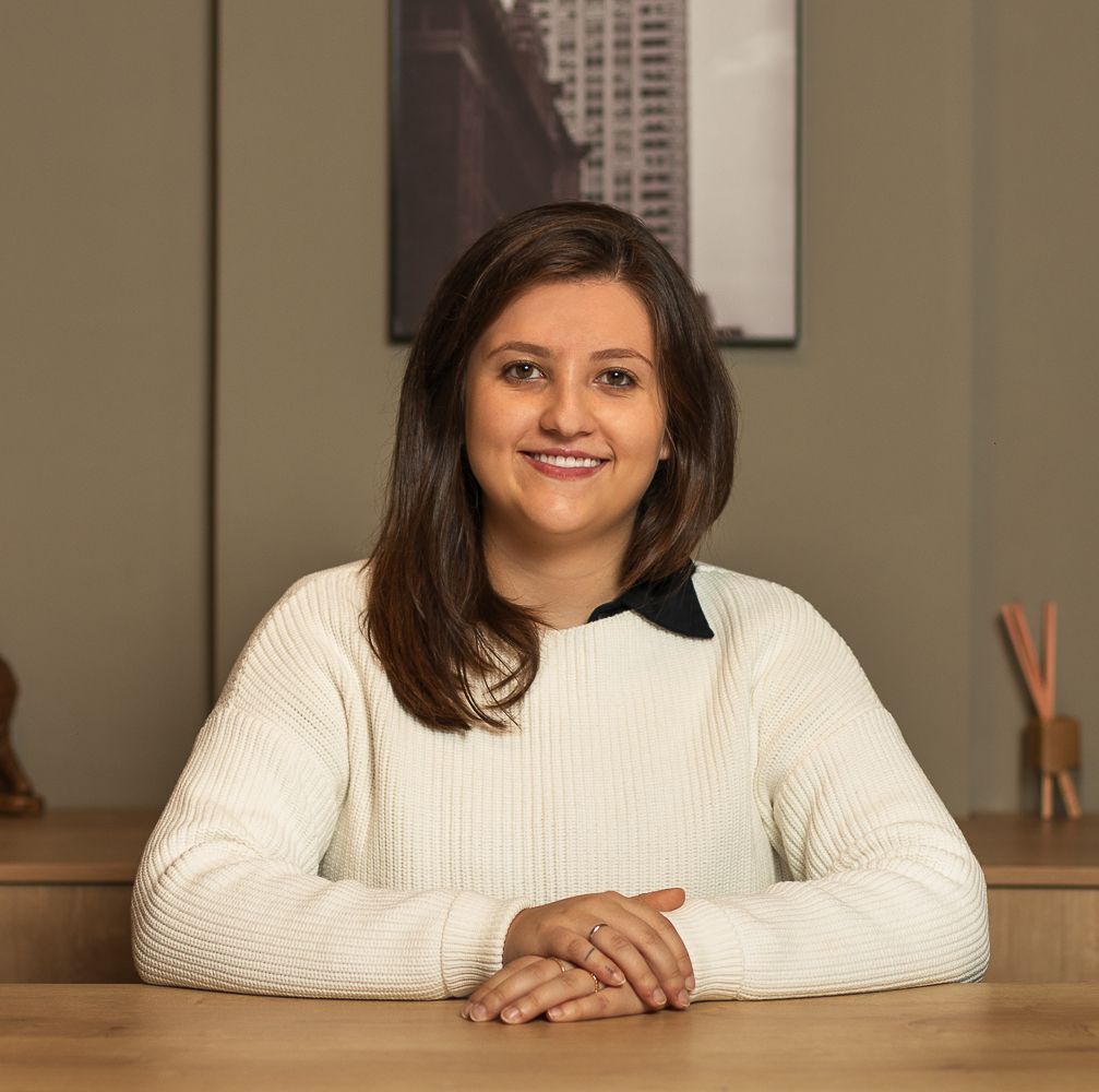 Uma mulher de suéter branco sorri enquanto está sentada em uma mesa, com obras de arte e objetos de decoração atrás dela.