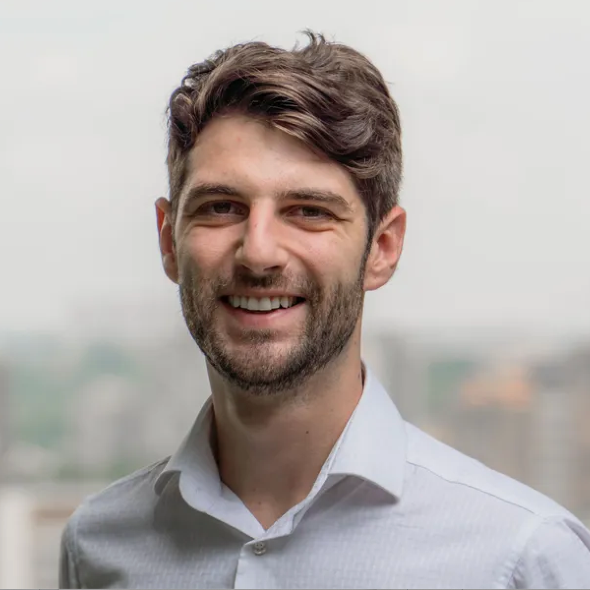 Homem de cabelos escuros e barba por fazer sorri amplamente, vestindo uma camisa branca de colarinho.