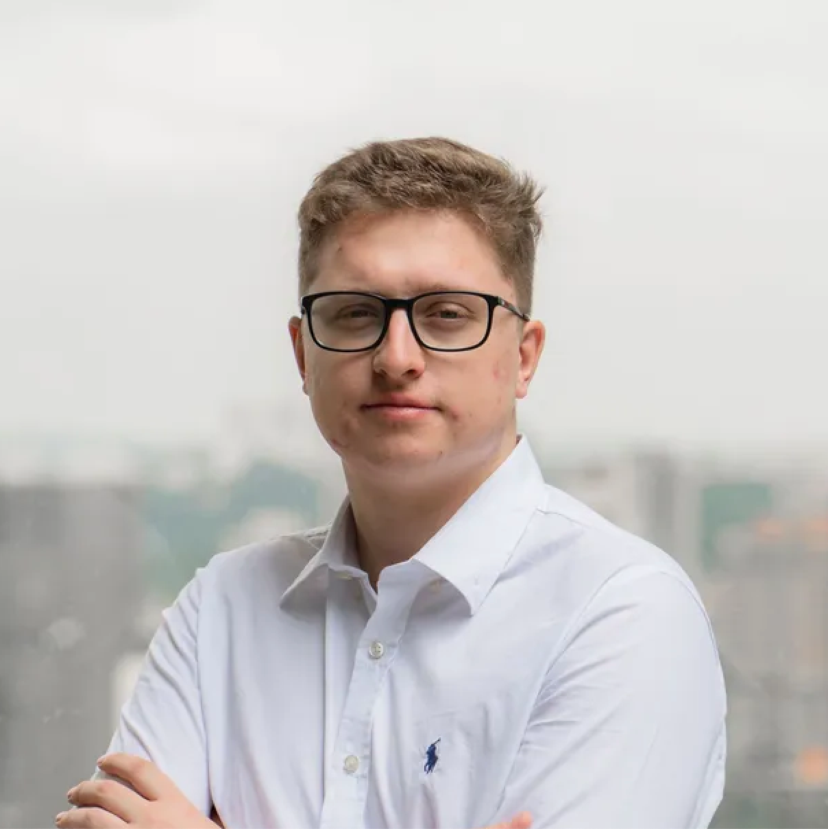 Homem de óculos e camisa branca, braços cruzados, olhando para a câmera tendo como pano de fundo uma paisagem urbana.