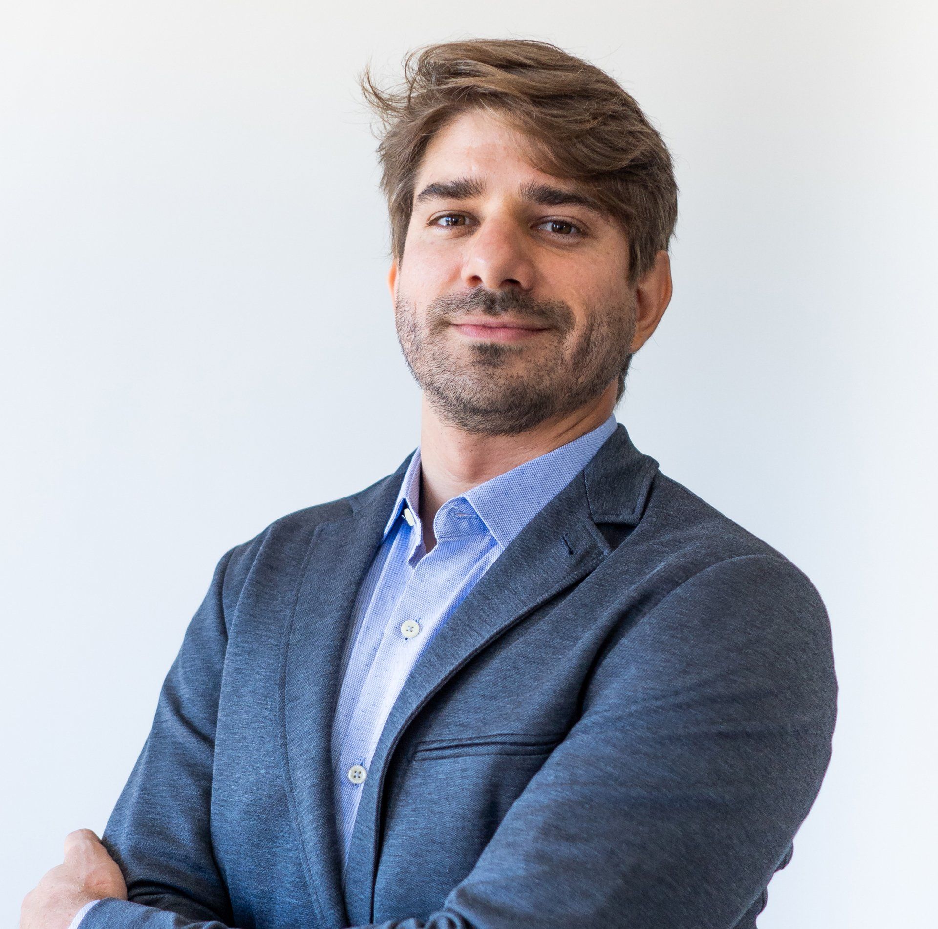 Homem barbudo, vestindo camisa azul e blazer cinza, com os braços cruzados e sorrindo.