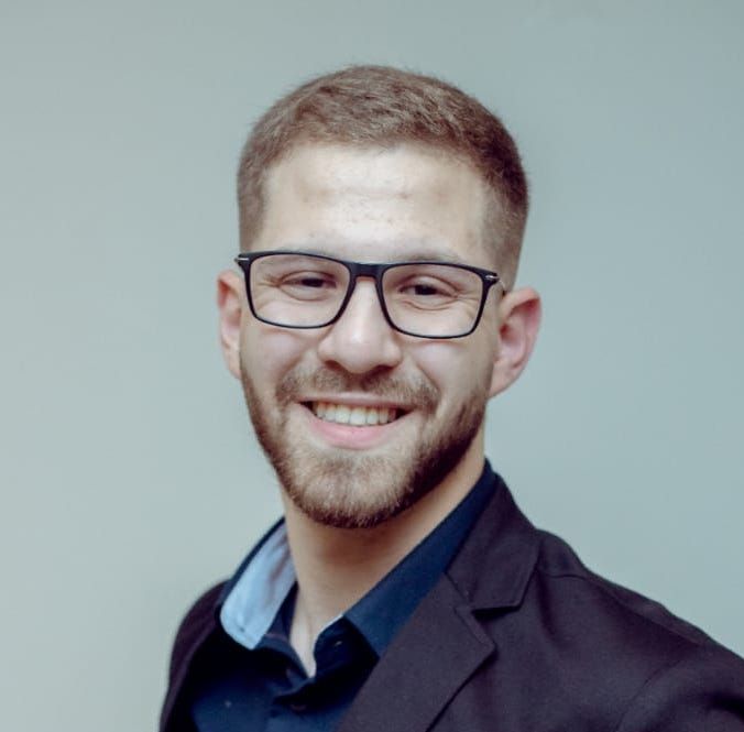 Homem de óculos e barba, sorrindo, vestindo blazer e camisa de botões.
