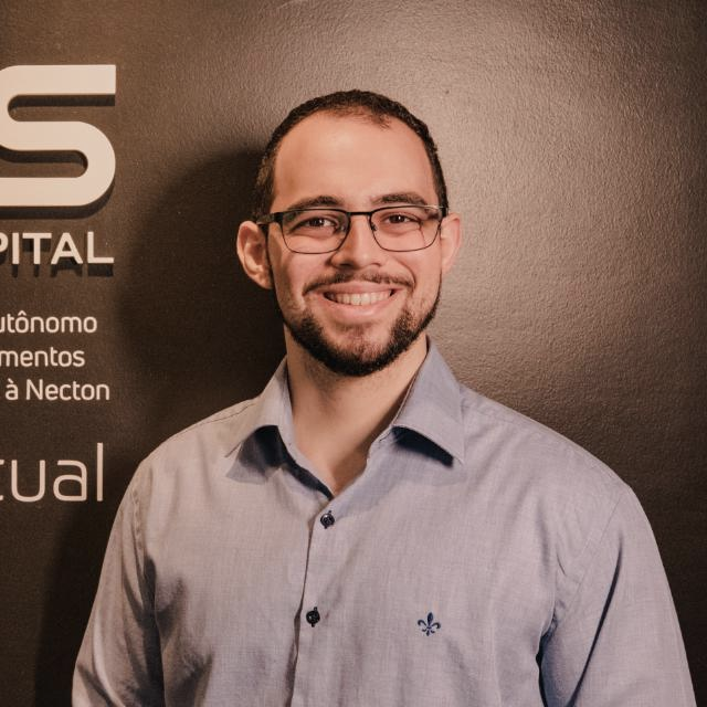 Homem de óculos, camisa azul-clara, sorrindo em frente a uma parede preta com texto branco.