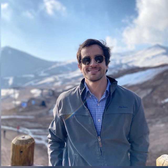 Homem de óculos escuros e jaqueta sorri em um cenário de montanha nevada.