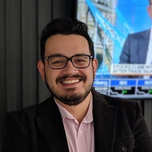 Homem de óculos sorrindo, vestindo uma camisa rosa e um blazer preto, em frente a uma tela que exibe notícias financeiras.