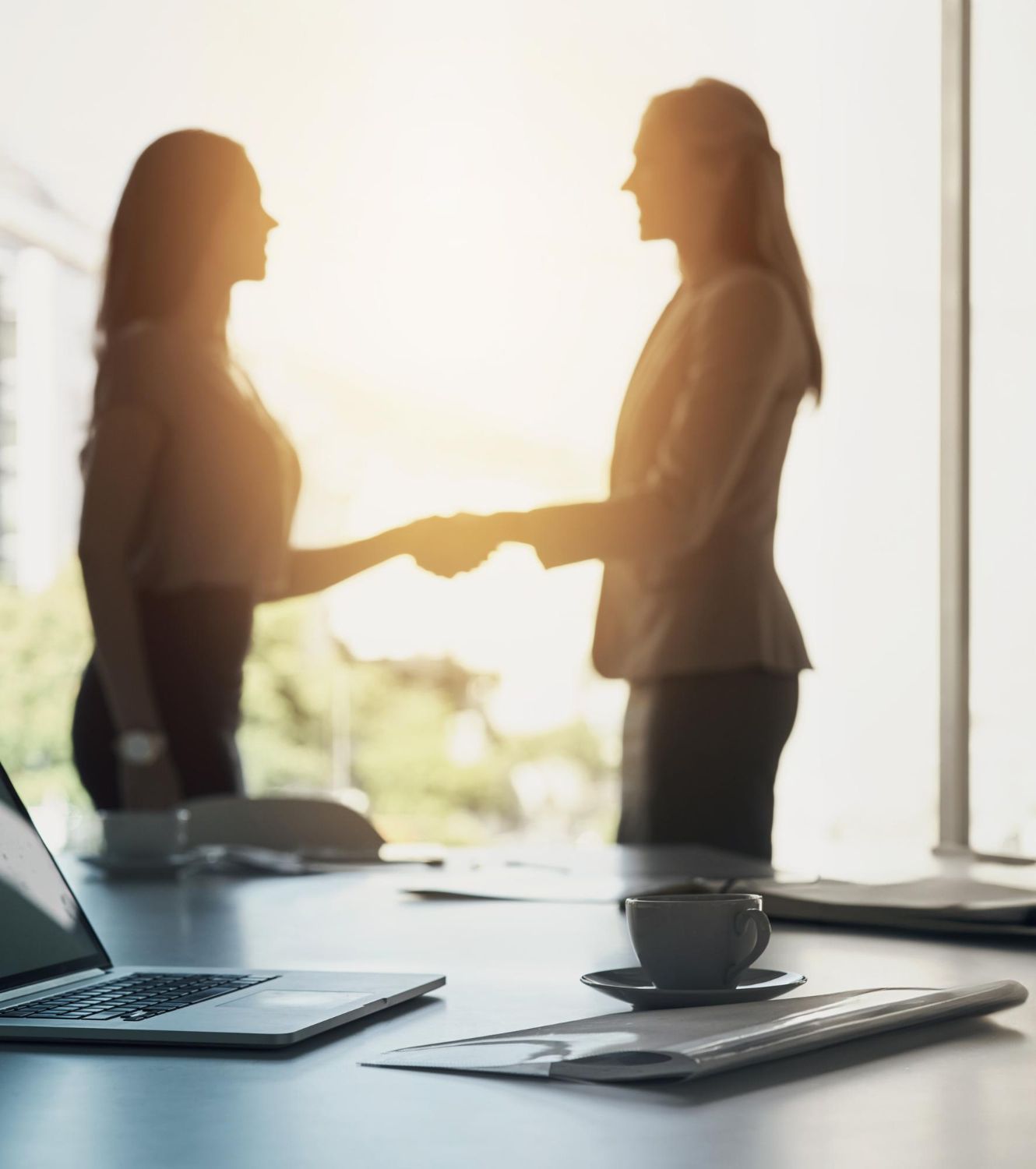 Two women in business attire shaking hands in front of a bright window; a laptop and coffee on the desk.