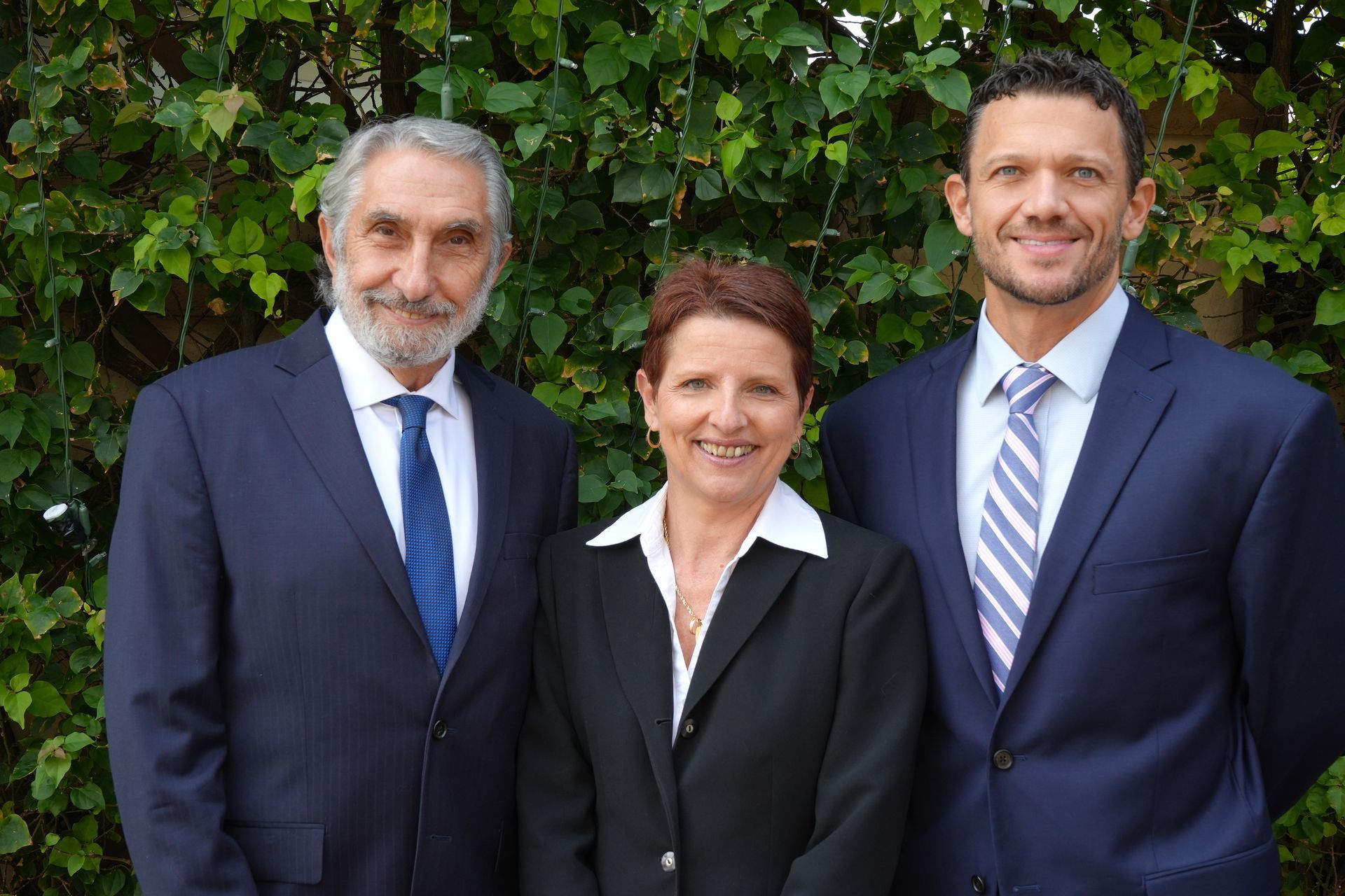 Three people in formal business attire pose smiling in front of a green leafy hedge.