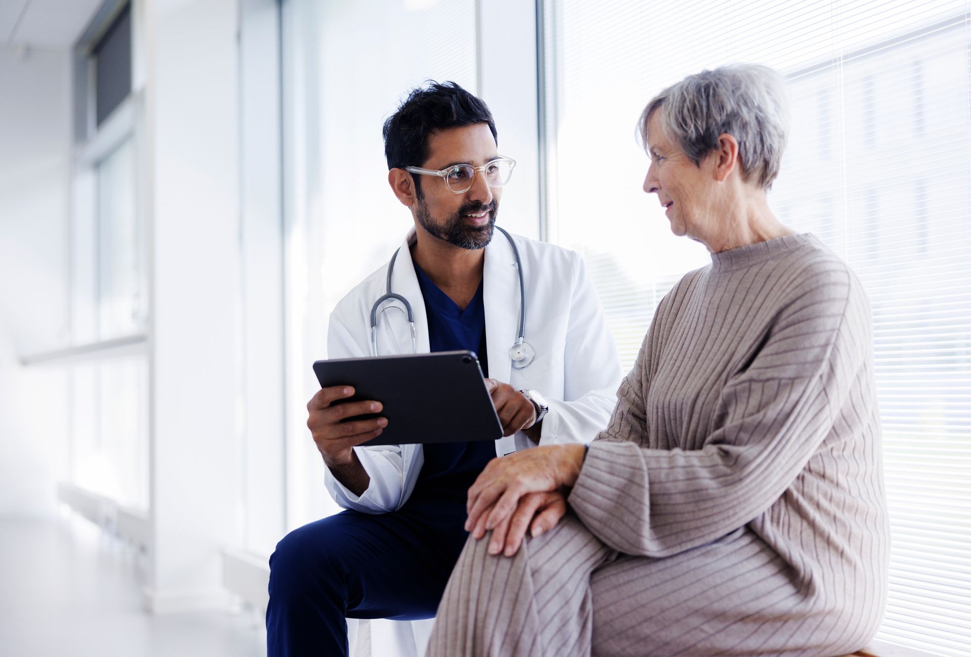 A doctor is talking to an elderly woman while holding a tablet.