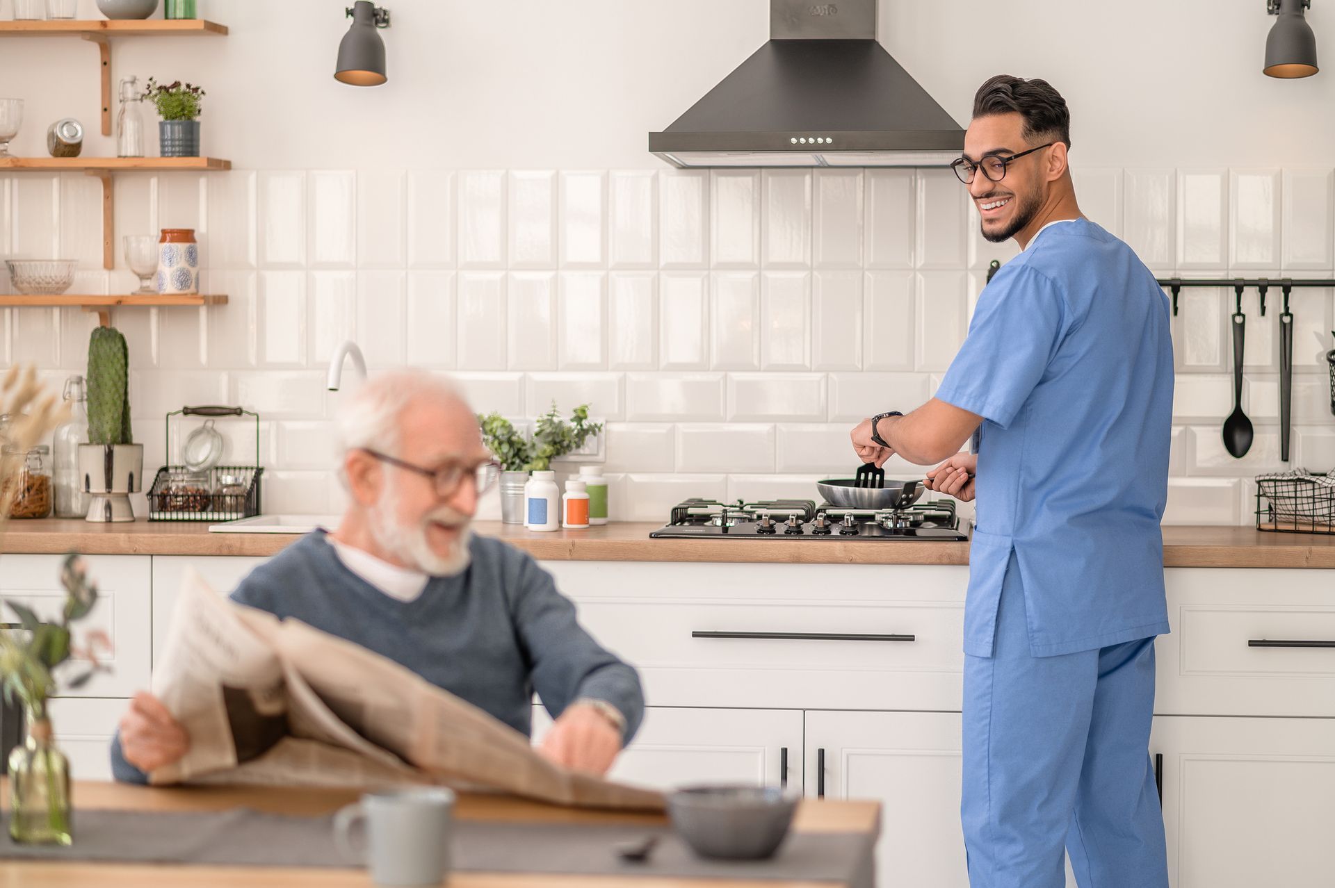 An elderly man is reading a newspaper while a nurse prepares food in the kitchen.