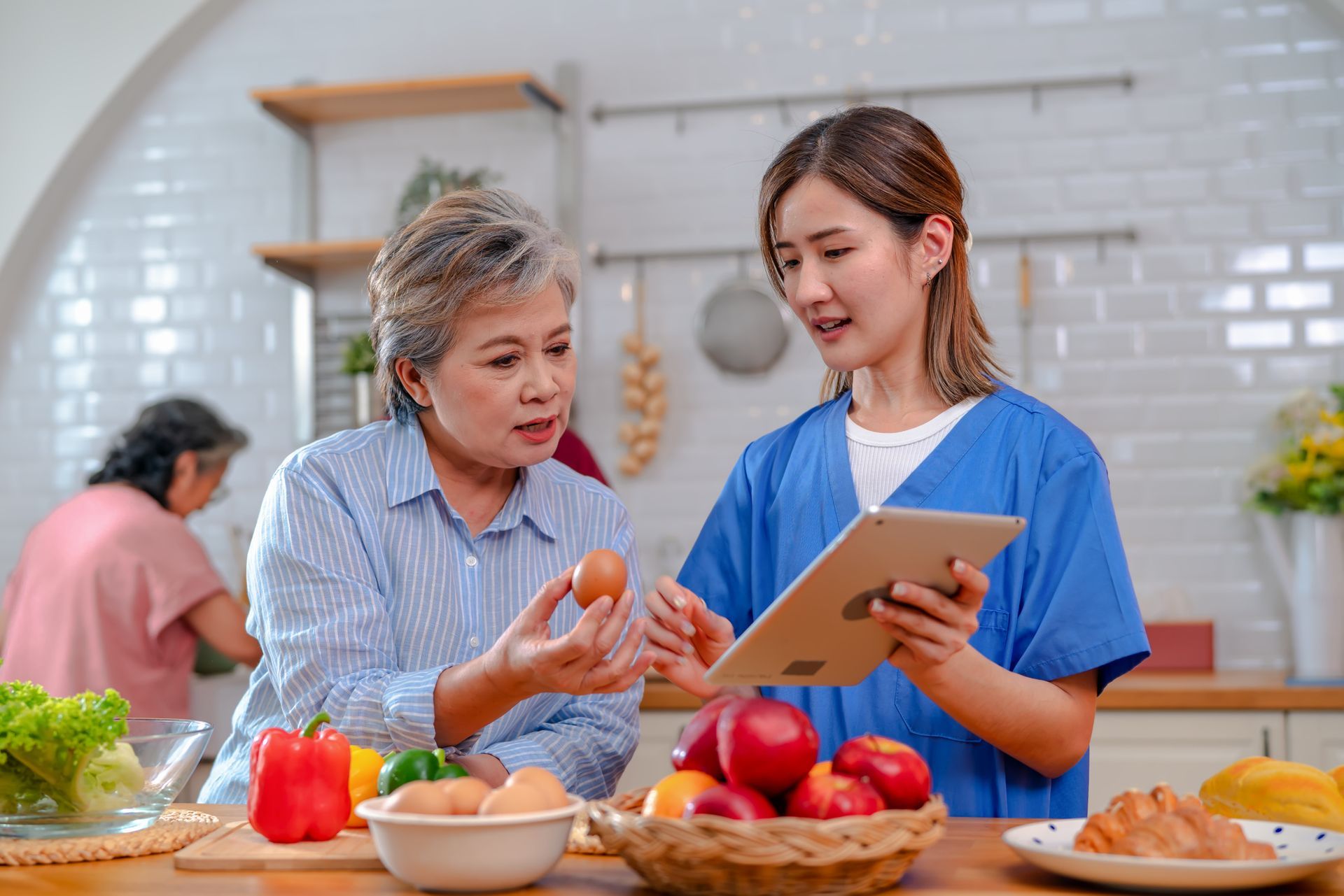 A nurse is talking to an elderly woman in a kitchen while looking at a tablet.