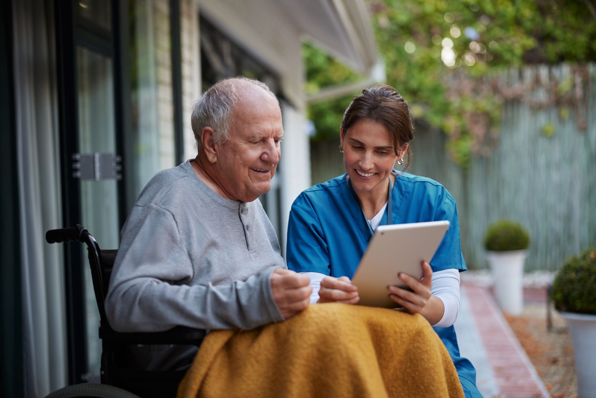 An elderly man in a wheelchair is sitting next to a nurse looking at a tablet.