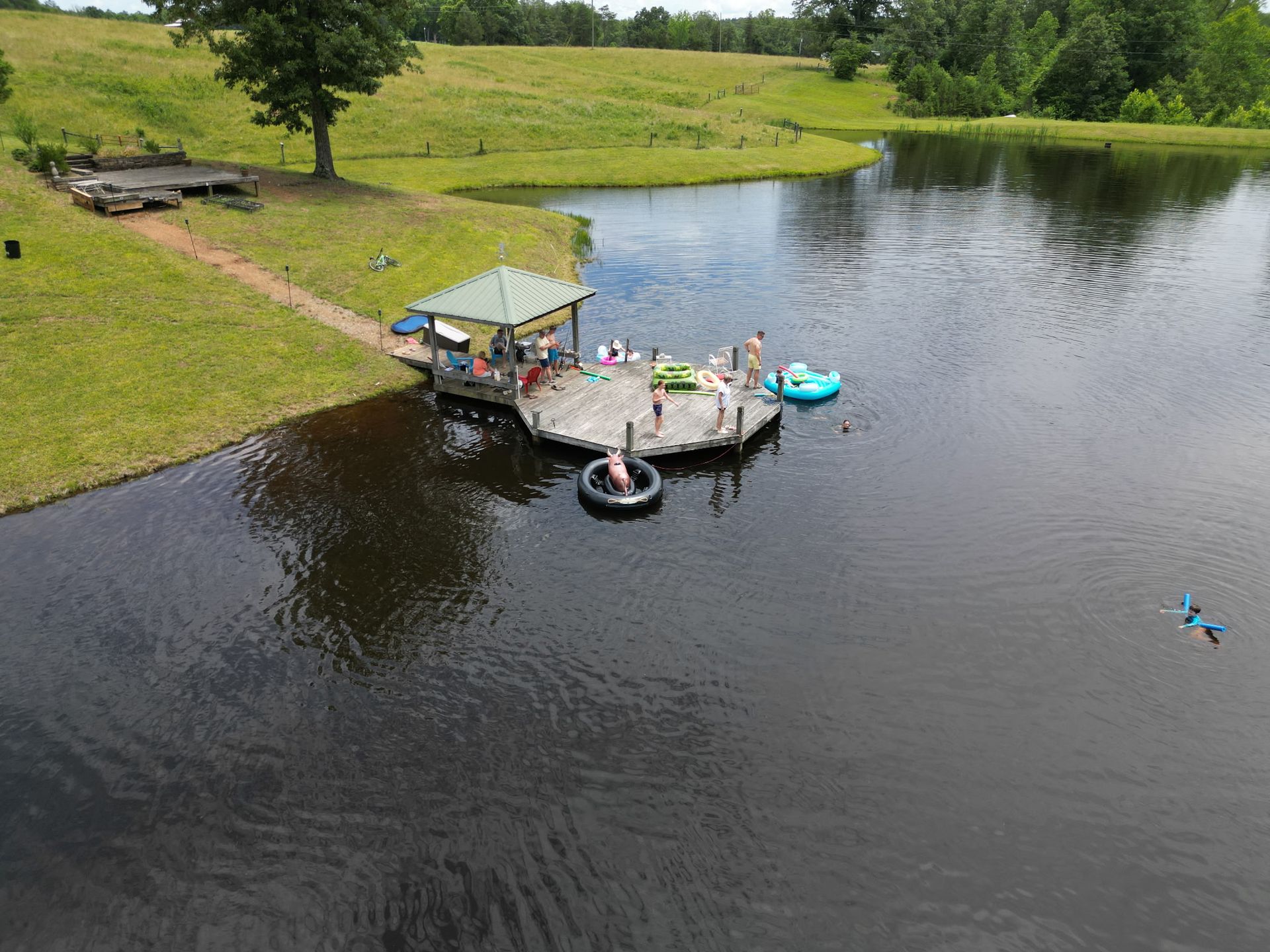 People on a dock, some in the water, at a pond. Green grass and trees in the background.