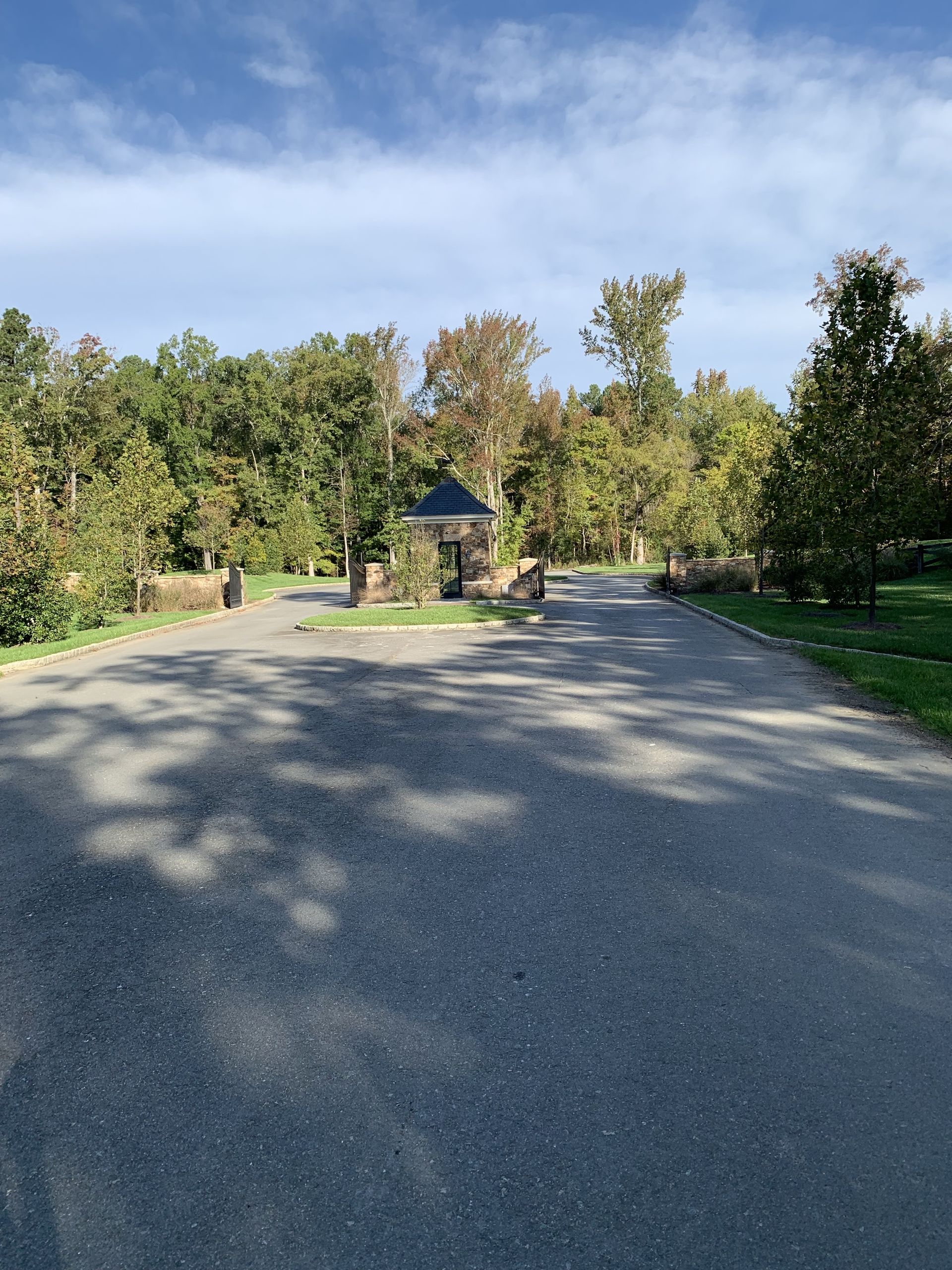 Asphalt road leading to a gated entrance surrounded by trees under a partly cloudy sky.