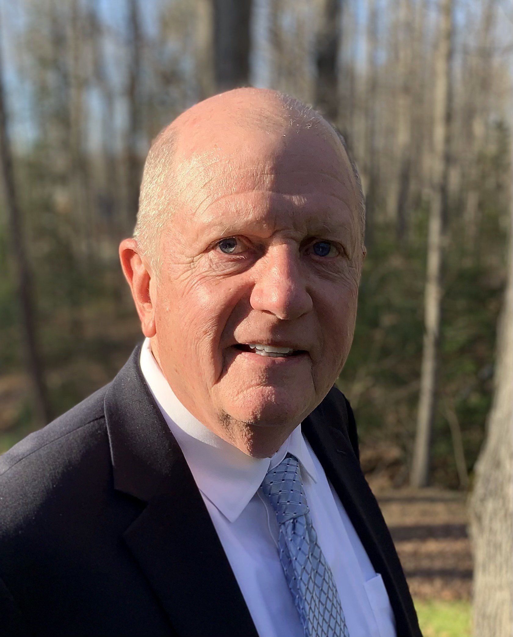 Bald, older man in a suit smiles outdoors.