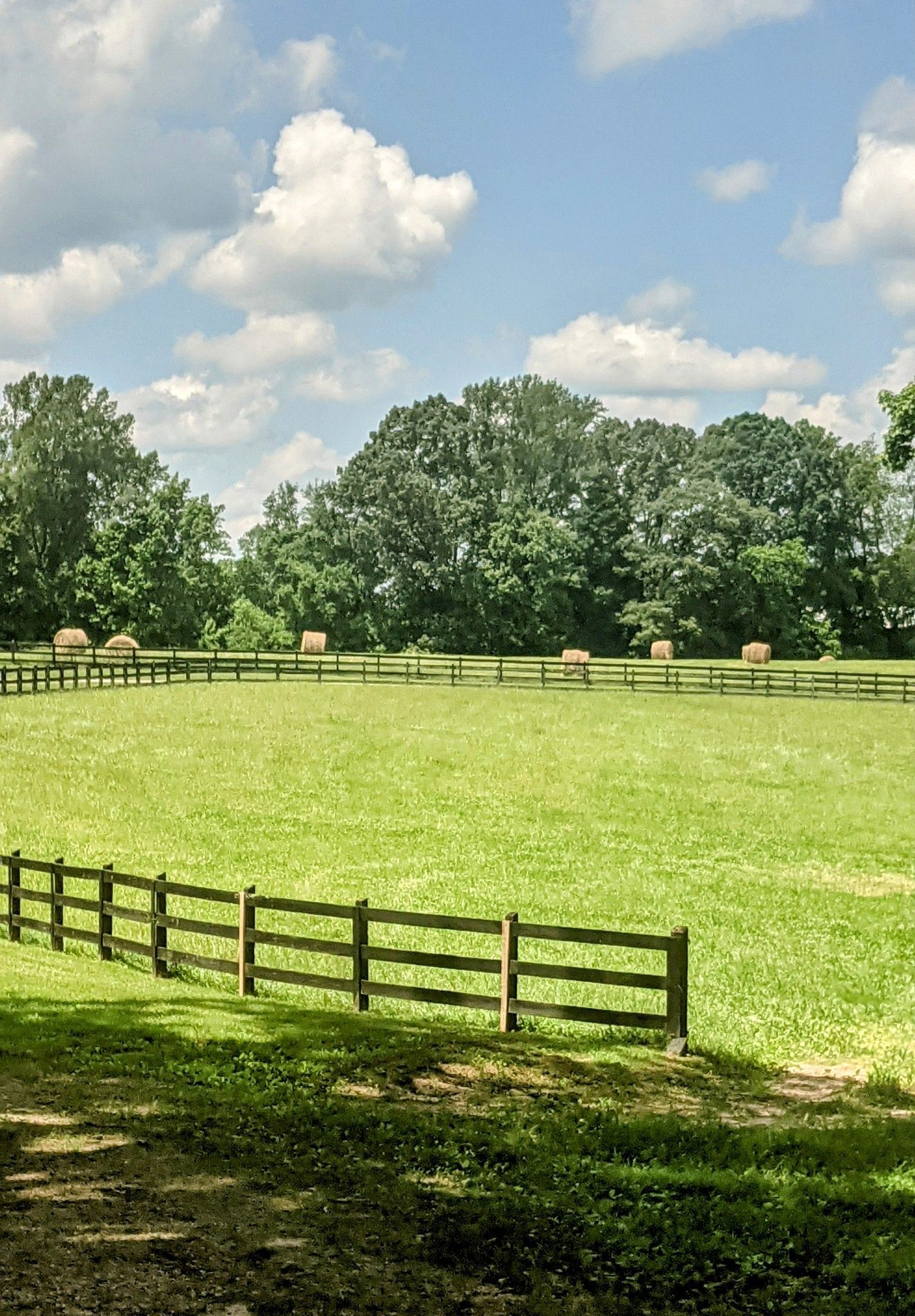 Grassy pasture with a wooden fence, hay bales, and trees under a partly cloudy sky.