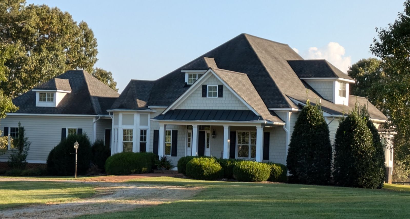 Large, white house with a dark gray roof, front porch, and dormers, set on a green lawn under a blue sky.