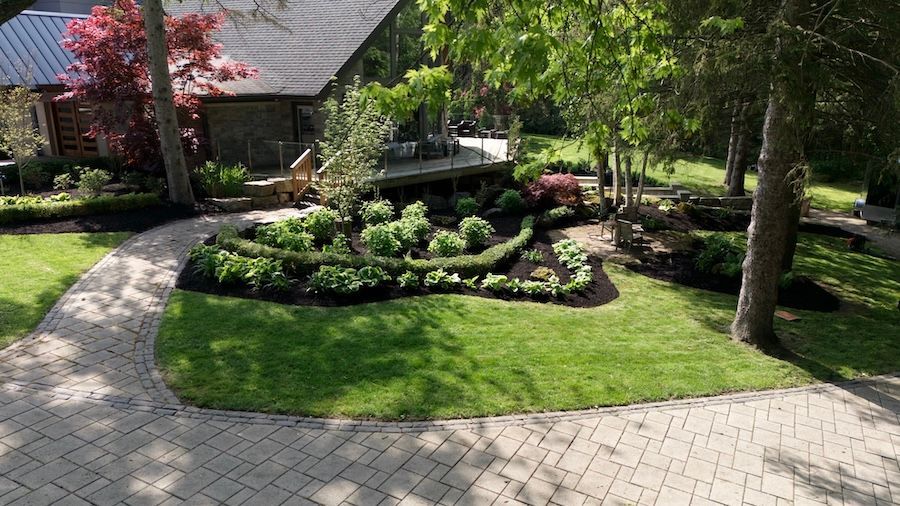 A brick walkway leads to a landscaped yard with curved garden beds, shrubs, and a house with a wooden deck in the back.