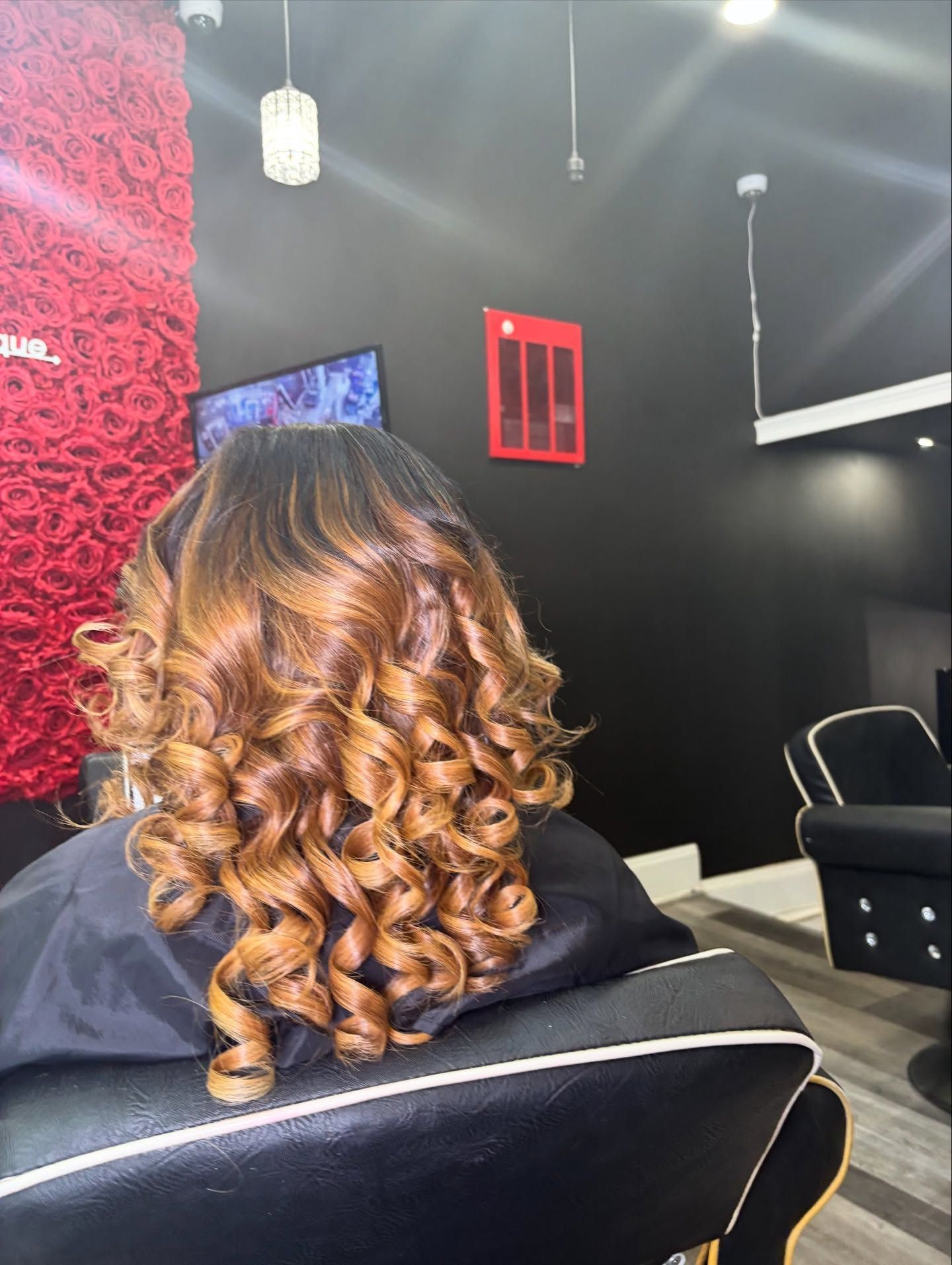 A person with highlighted, curled brown hair sits in a salon chair in front of a red rose wall decor.