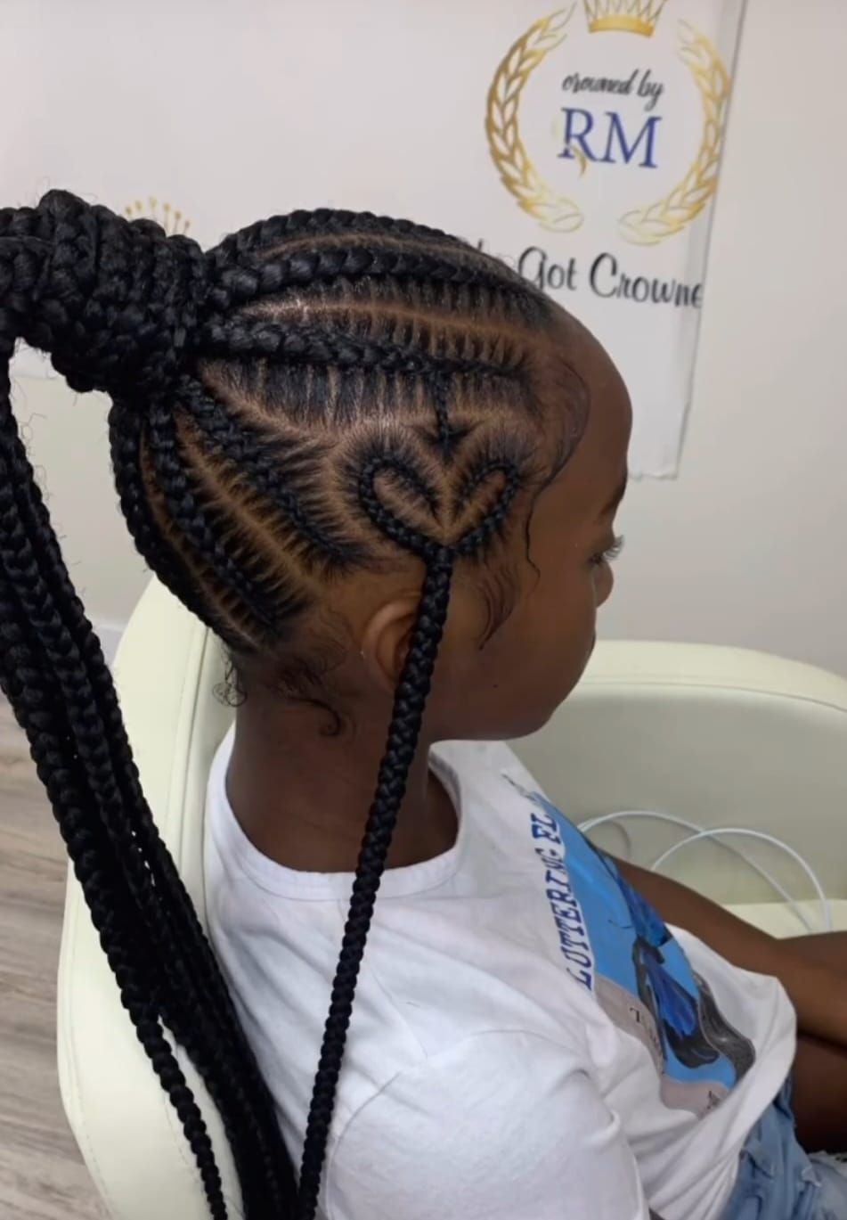 A person’s head featuring intricate cornrow braids styled into a high ponytail, with a heart-shaped design on the side.