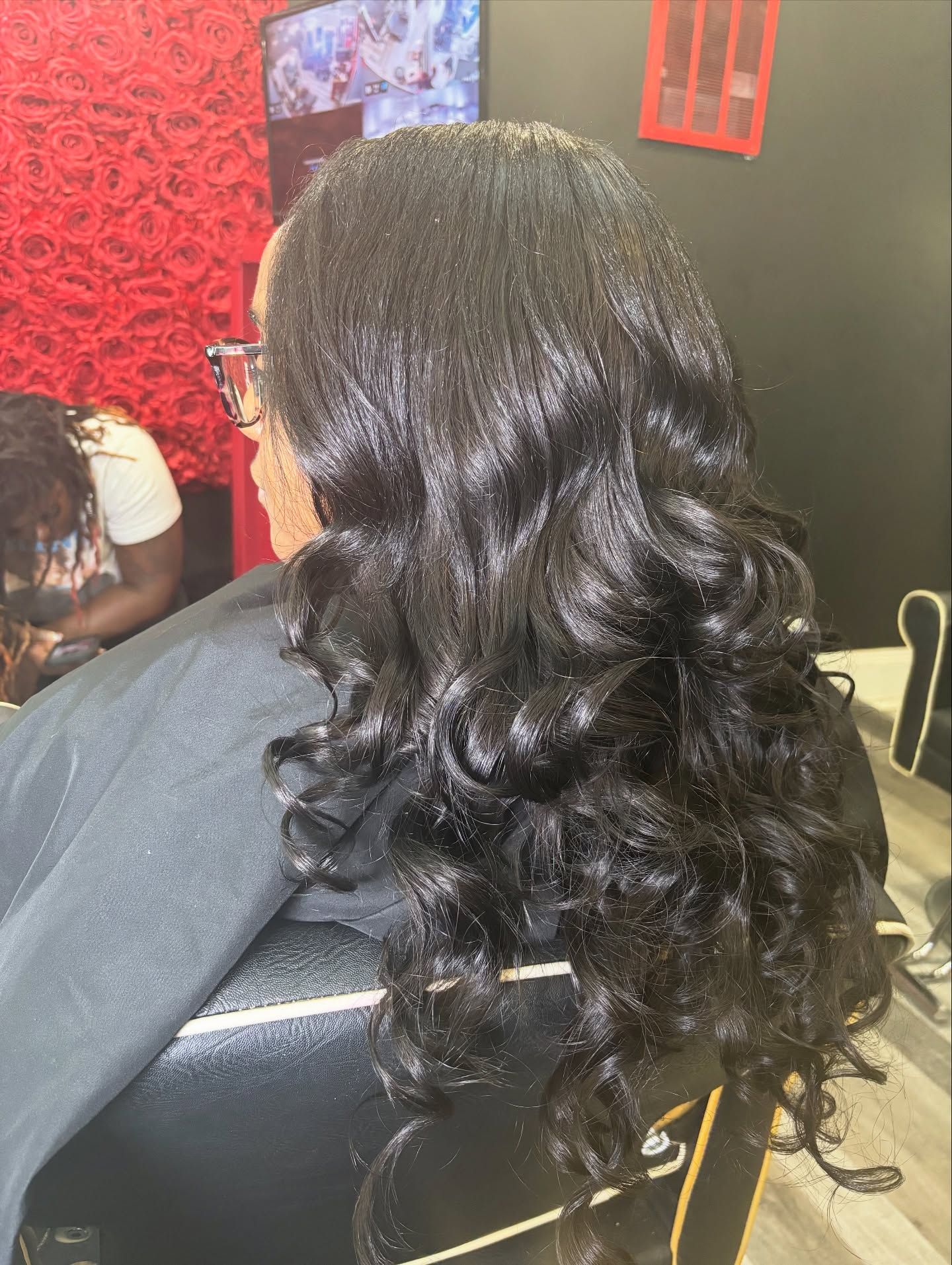 A person with long, dark, wavy hair styled in a salon setting, wearing a black cape.