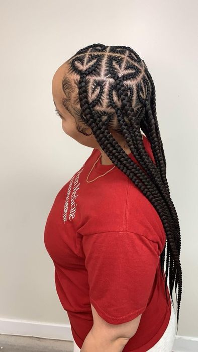 A person wears their dark hair styled in long braids with intricate, heart-shaped partings along the scalp.