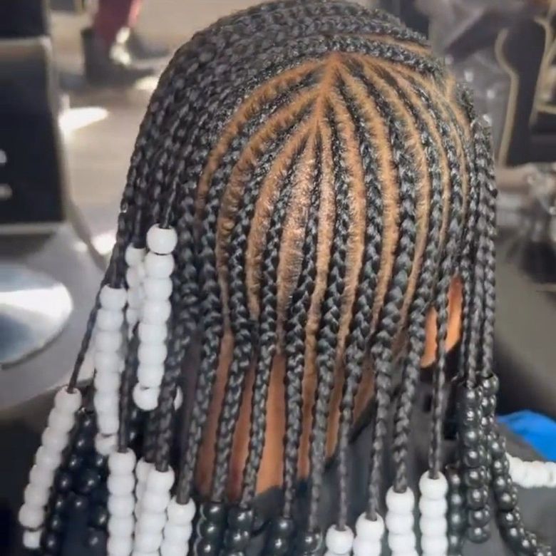 A high-angle close-up of a person with cornrow braids parted in a starburst pattern, accented with black and white beads.