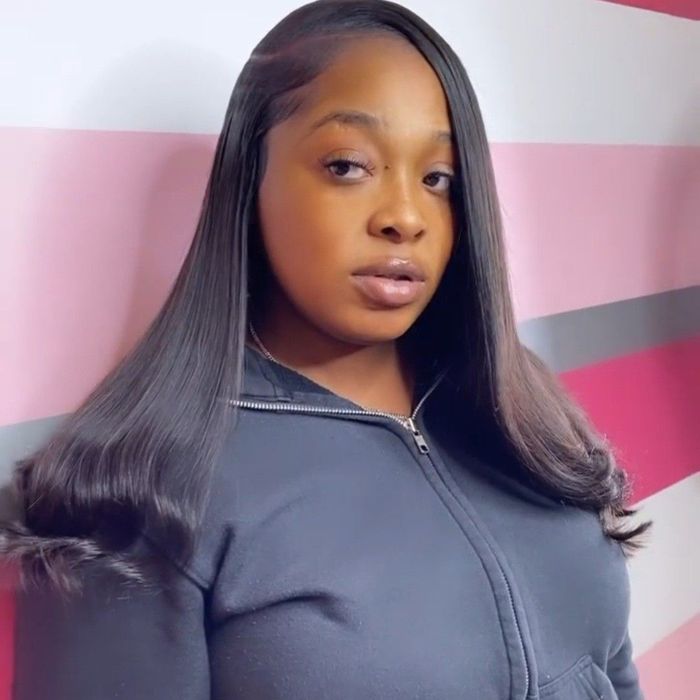 A person with long, sleek, black hair wearing a dark zip-up hoodie, posed against a pink and white striped wall.