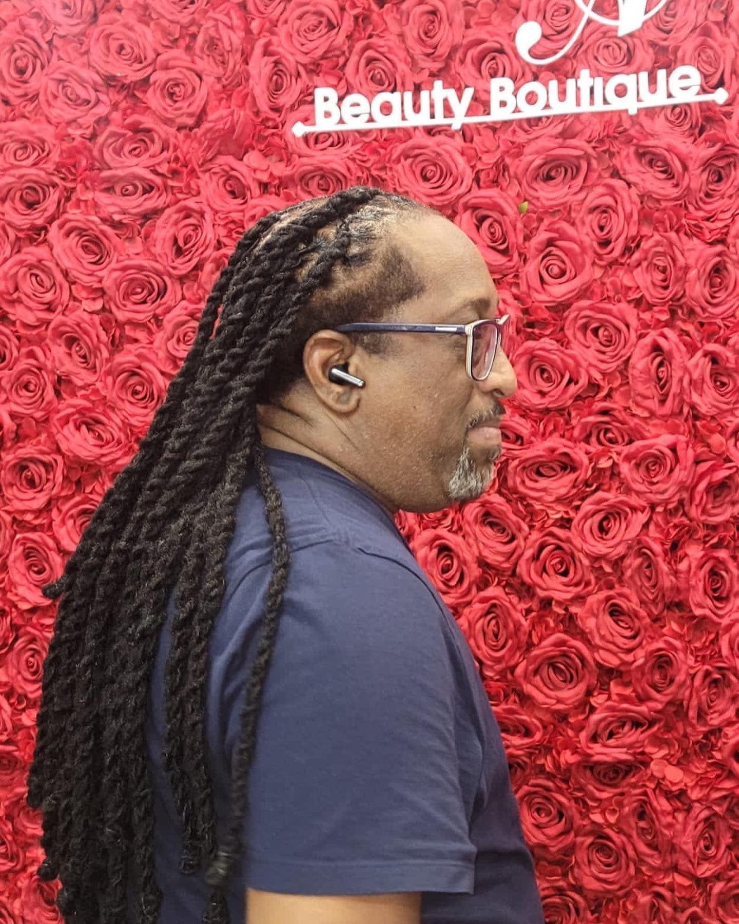 A person with long, dark, twisted hair wearing glasses and an earbud, posed in profile against a red rose wall backdrop.