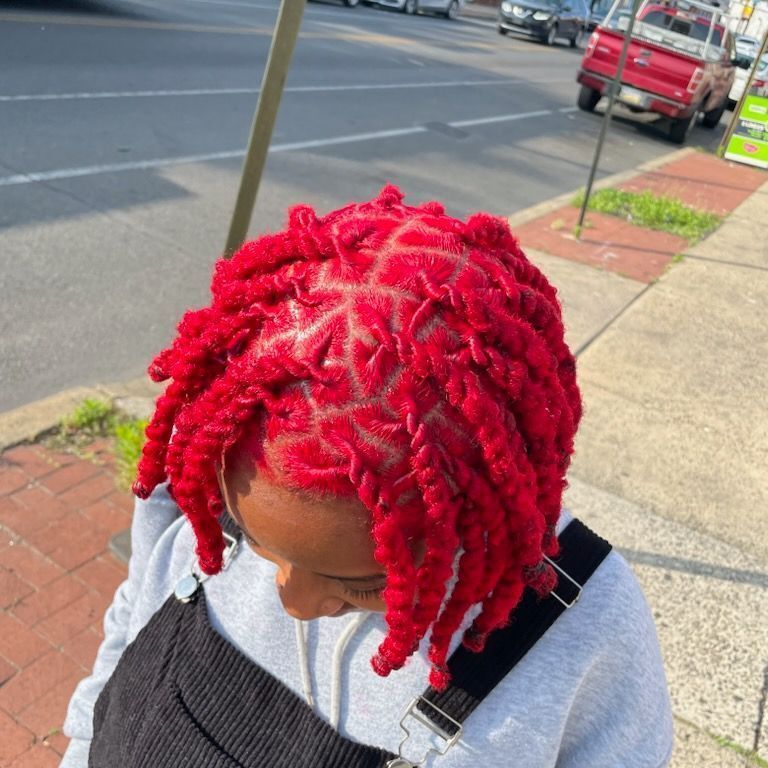 A person with vibrant, bright red two-strand twists sectioned into small triangles on a city sidewalk.