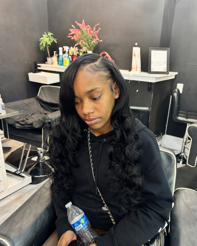 A person with long, wavy dark hair and a side part sits in a salon chair, holding a water bottle.