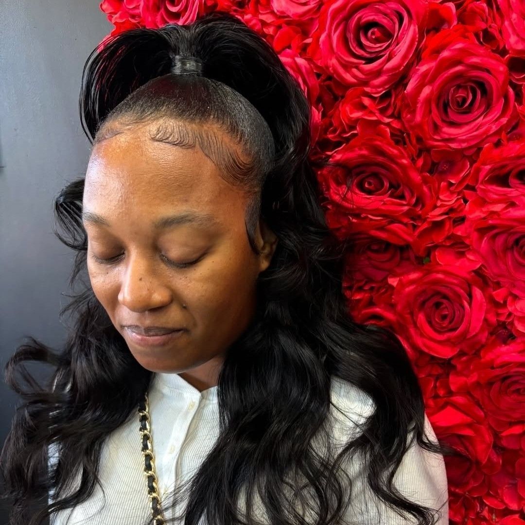 A person with dark, wavy hair styled in a half-up, half-down look stands against a backdrop of vibrant red roses.