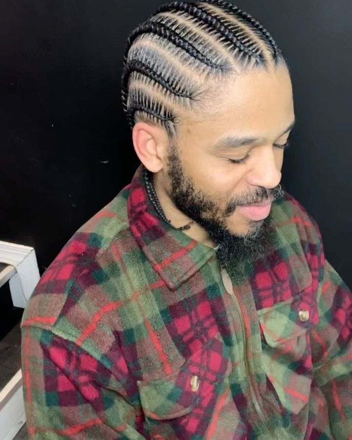 A man with a groomed beard wears a red, green, and black plaid shirt, showcasing neat, straight-back cornrow braids.