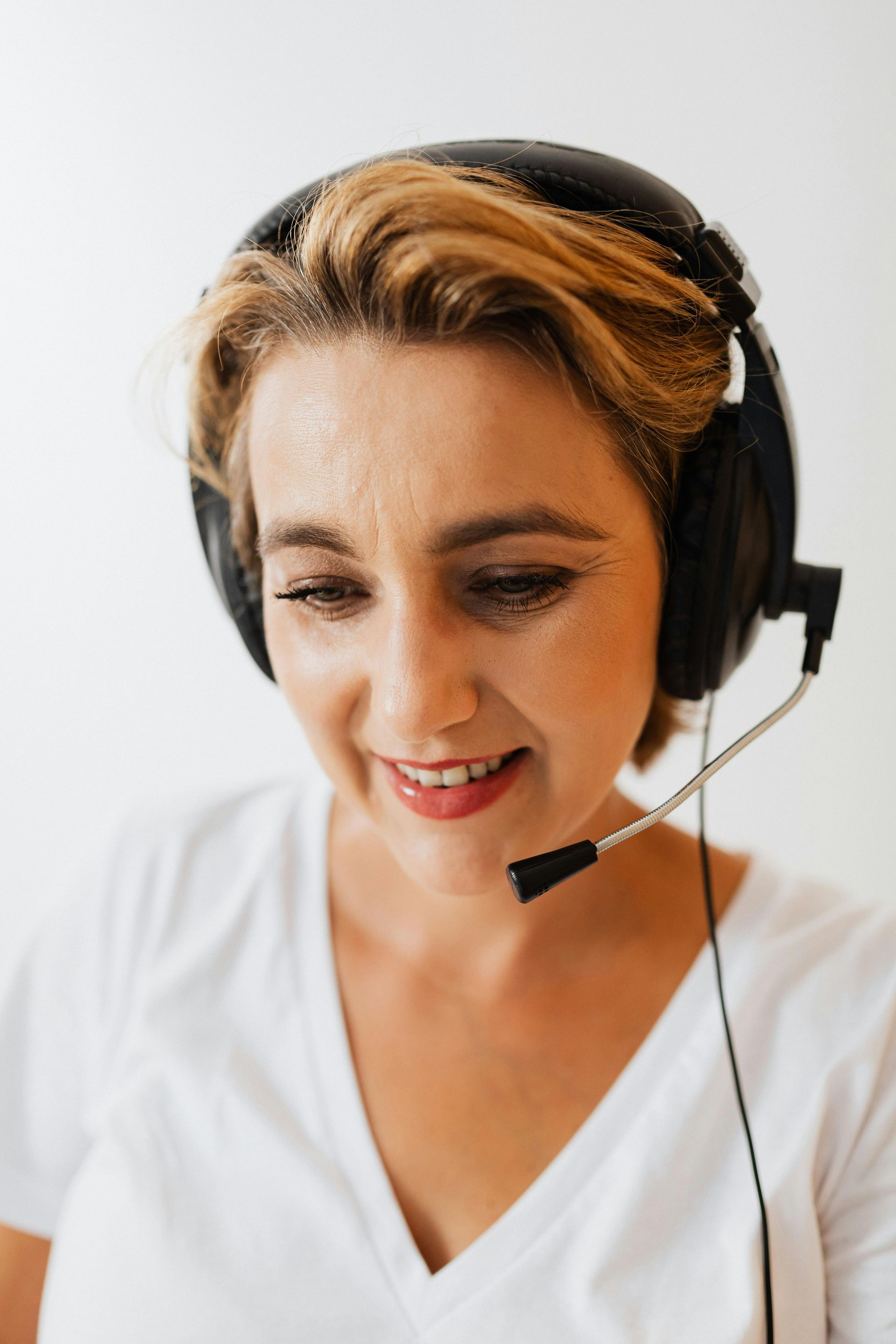 Mujer con auriculares, sonriendo mientras mira hacia abajo, camisa blanca sobre fondo blanco.