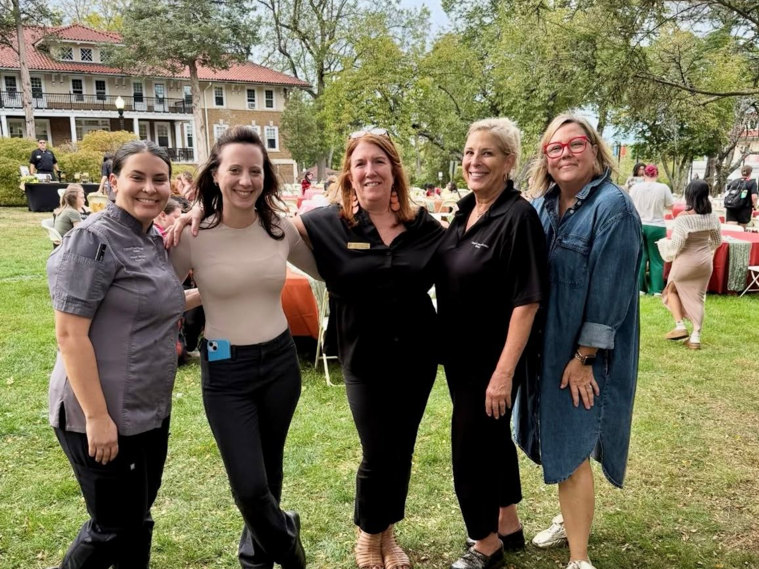 Five smiling people pose outdoors. One has arm around another. Buildings and event in background.