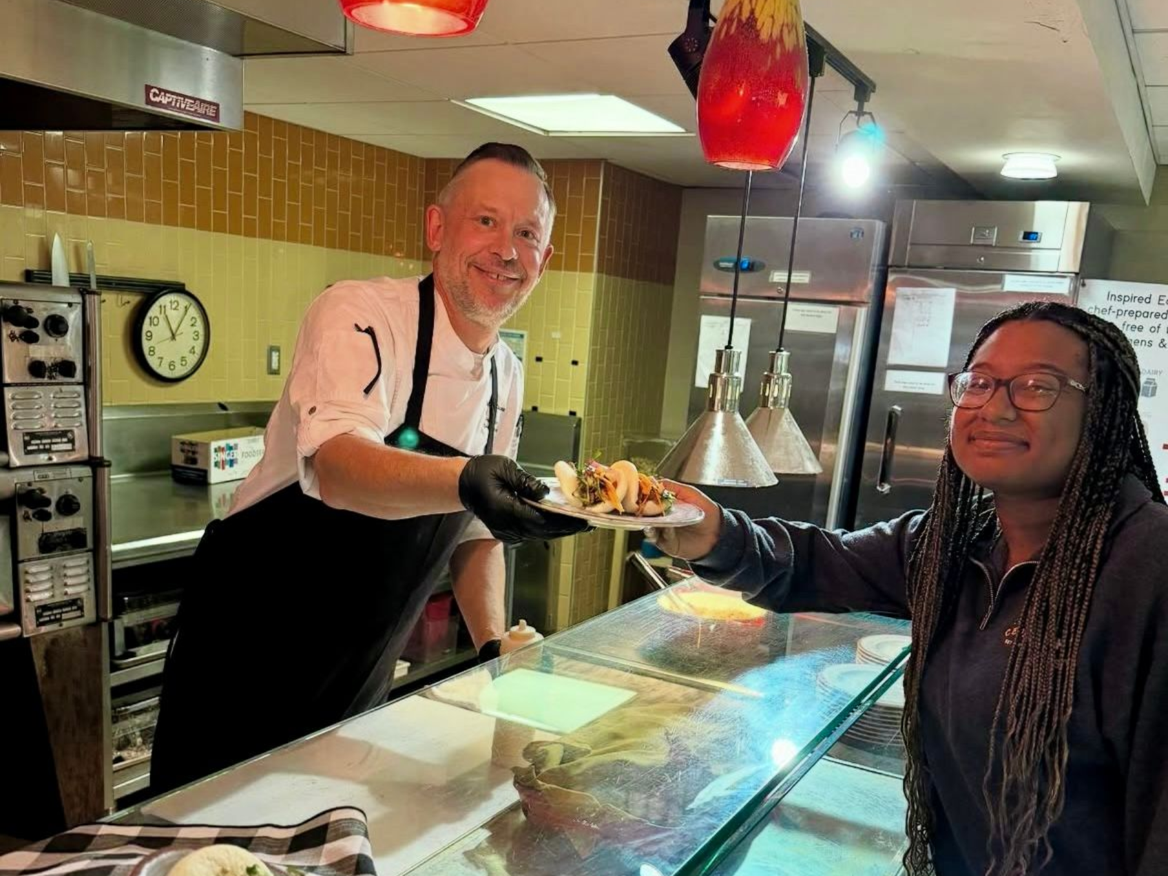 Chef hands tacos to a customer at a restaurant counter. Both smile, chef wears a black apron.