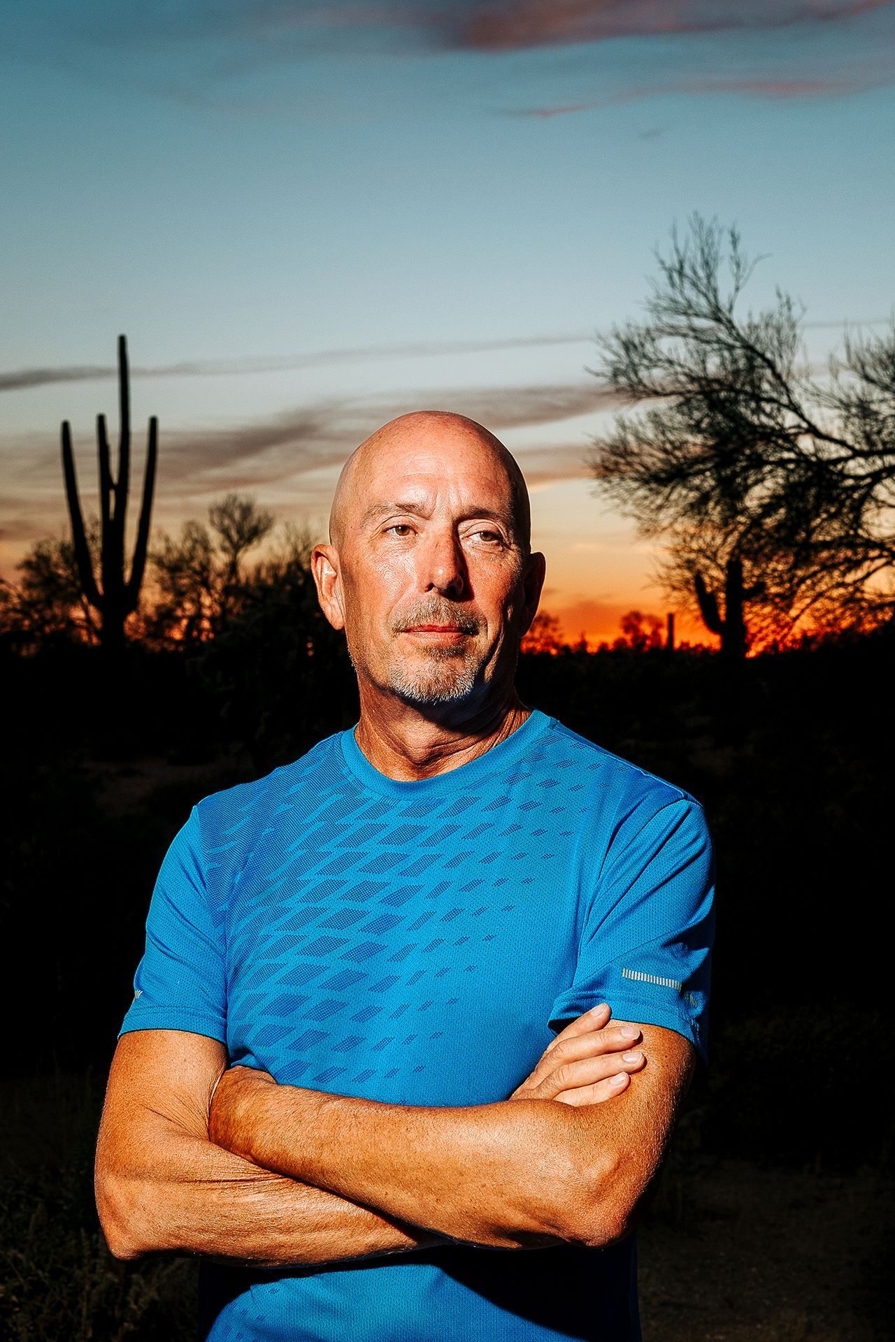A man in a blue shirt is standing in the desert with his arms crossed.